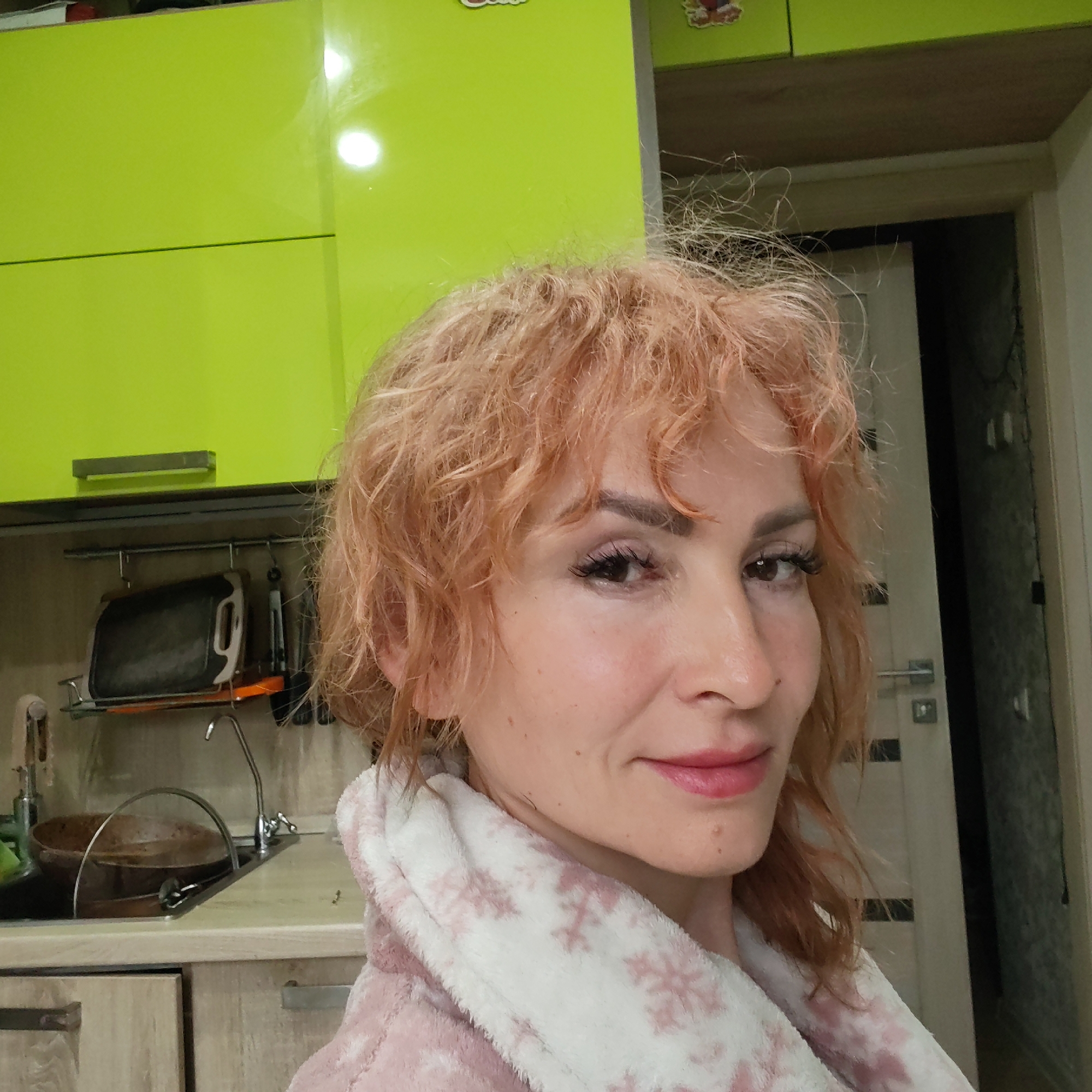 A woman is in a kitchen in Chaikovsky, Russia. She has short, wavy, reddish-blonde hair and is wearing a pink and white bathrobe with snowflake patterns. Behind her are green kitchen cabinets, a dish rack over a sink, and a partially open doorway.
