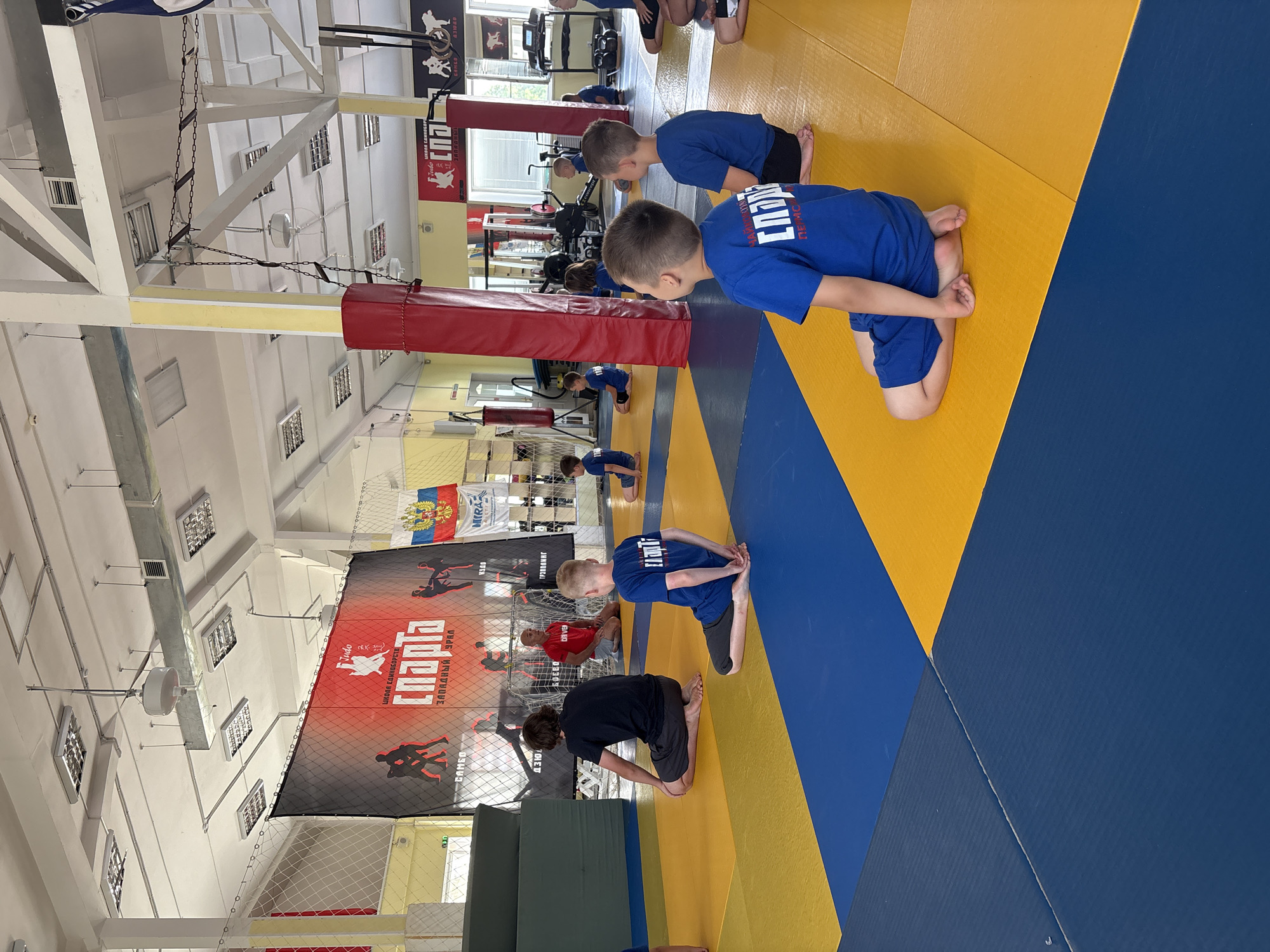In an indoor martial arts training facility in Chaikovsky, Russia, a group of children and a few adults are sitting on blue and yellow mats. The children, mostly boys, are wearing blue t-shirts with Cyrillic lettering that appears to say "СПАРТА" (SPARTA) and blue shorts or athletic pants. They are seated in a formal or attentive posture, facing a presumed instructor who is not clearly visible in the foreground. The environment includes large banners on the wall depicting martial arts imagery, Russian flags, and sports equipment such as punching bags and weight machines visible in the background. The lighting appears to be from overhead fluorescent fixtures. The overall scene suggests a martial arts class or practice session for young students.