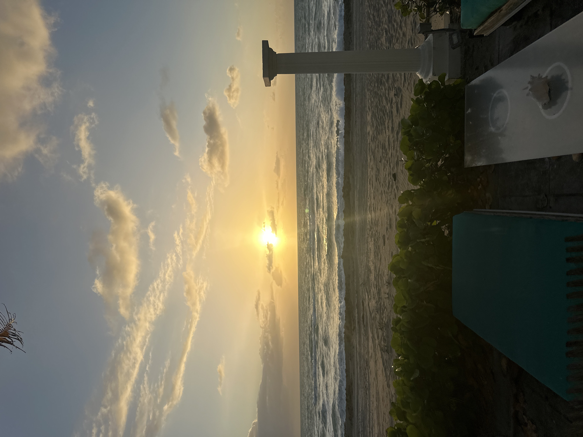 The image depicts a beach scene during sunset in Hallandale Beach, Florida, United States. The sun is near the horizon, casting a golden glow on the sky and ocean. The sky features clouds in various forms. Waves are breaking on the beach, and vegetation lines the edge of the shore. A white column structure stands in the foreground near some furniture. The image is oriented vertically.