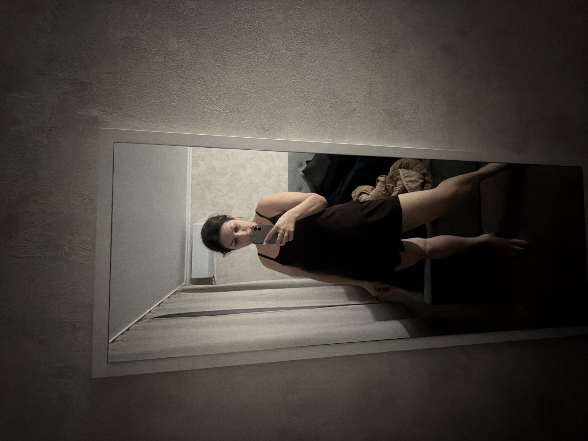 A person stands in front of a full-length mirror, holding a smartphone to take a picture of their reflection. They are wearing a dark-colored slip dress. The mirror is positioned in a dimly lit room with textured walls. In the reflection, a bed and some draped fabric are visible in the background. The person's legs and feet are partially visible at the bottom of the frame. The scene appears to be indoors, likely in a residential setting, and there is no indication that it is located in Sochi, Russia.