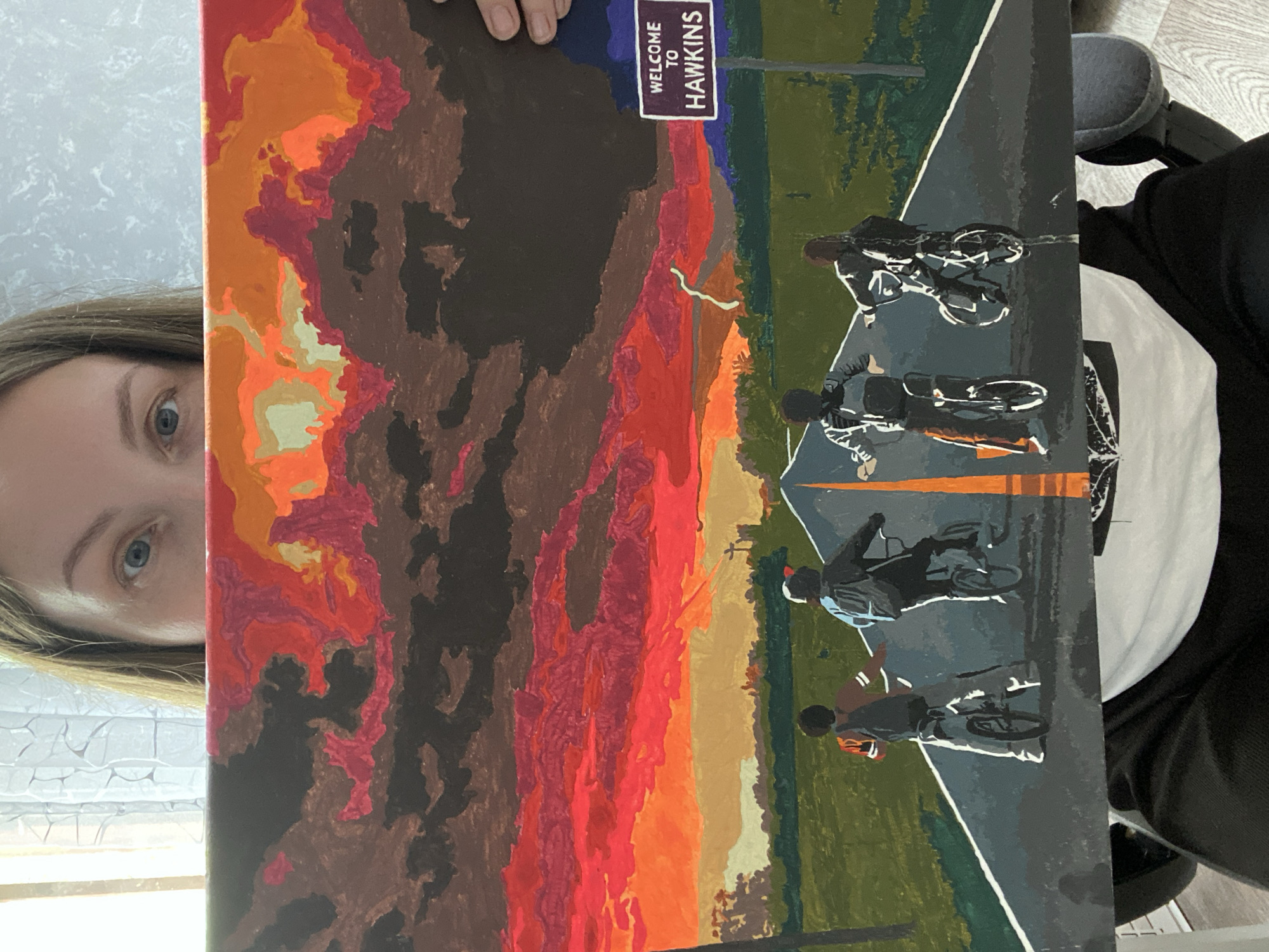 The image shows a person holding a painting. The painting depicts four individuals on bicycles riding down a road towards a "Welcome to Hawkins" sign. The sky in the painting is rendered with dark, stormy colors including brown, red, orange, and yellow. The road is grey with a white stripe and orange markings. Green vegetation borders the road. The painting appears to be inspired by the television show "Stranger Things." The location is not specified in the image.