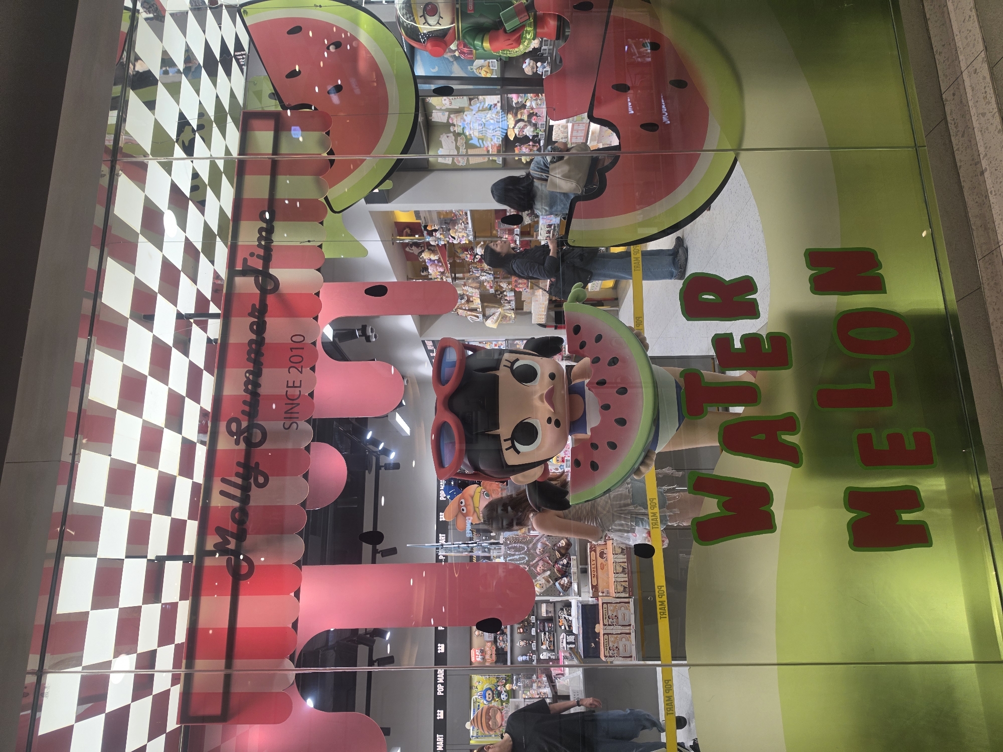 This is an indoor shot of a retail display window in Bangkok, Thailand, advertising a line of "Molly Summer Jimo" figurines since 2010. The storefront appears to be a POP MART location. The display features large, colorful, and whimsical graphics related to watermelon, including slices, patterns, and the words "WATER MELON" in large, bright green and red letters. There's a cartoon figurine with oversized red sunglasses holding a watermelon slice. The window reflects the store's interior, including shelves of merchandise and a few blurred people, presumably shoppers, are seen looking at the goods.