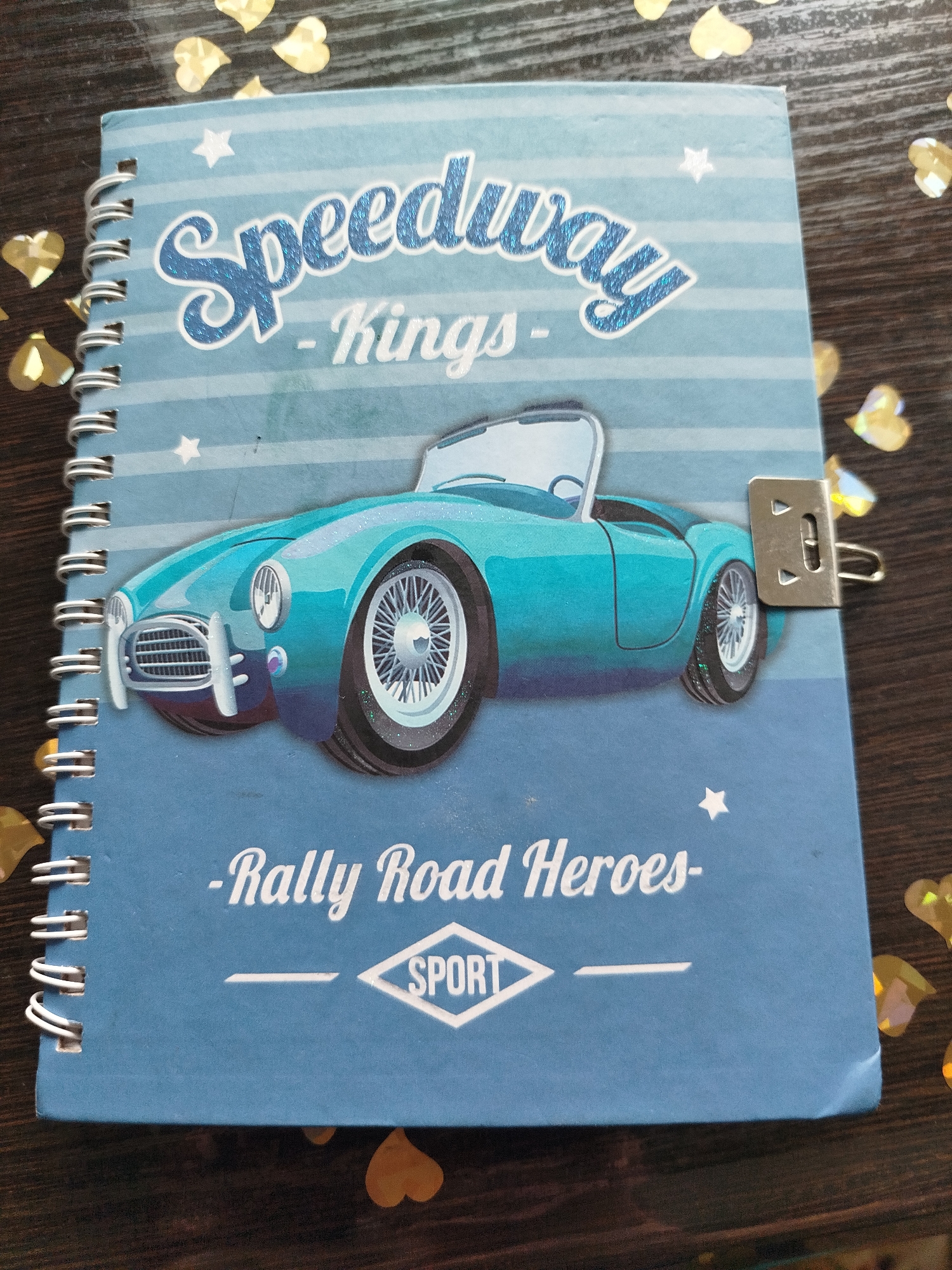 The media file is a top-down, medium shot of a blue spiral-bound notebook placed on a dark wooden surface. The notebook's cover features a graphic of a teal convertible sports car, likely a vintage model, with its front angled towards the viewer. The car is depicted with a glittery effect, especially on its wheels. The background of the notebook cover has light blue horizontal stripes and small white stars.
Prominently displayed at the top of the cover in large, stylized blue glitter text is "Speedway". Below this, in smaller white script, is "-Kings-". Towards the bottom of the cover, also in white script, is "-Rally Road Heroes-", with a diamond-shaped emblem containing the word "SPORT" to its right.
A small, silver metal clasp is attached to the right edge of the notebook, holding the pages together. The wooden surface beneath the notebook is partially visible and has a dark, grained texture. Scattered around the notebook, on the wooden surface, are several gold, heart-shaped confetti pieces.
The overall impression is that of a children's notebook or diary with a racing theme. There are no people visible, and the setting is a simple flat lay, suggesting an indoor or staged environment. The lighting appears to be neutral, and there are no indications of a specific time of day or weather conditions. The location context provided suggests a Belarusian setting, but no explicit visual cues within the image confirm this.