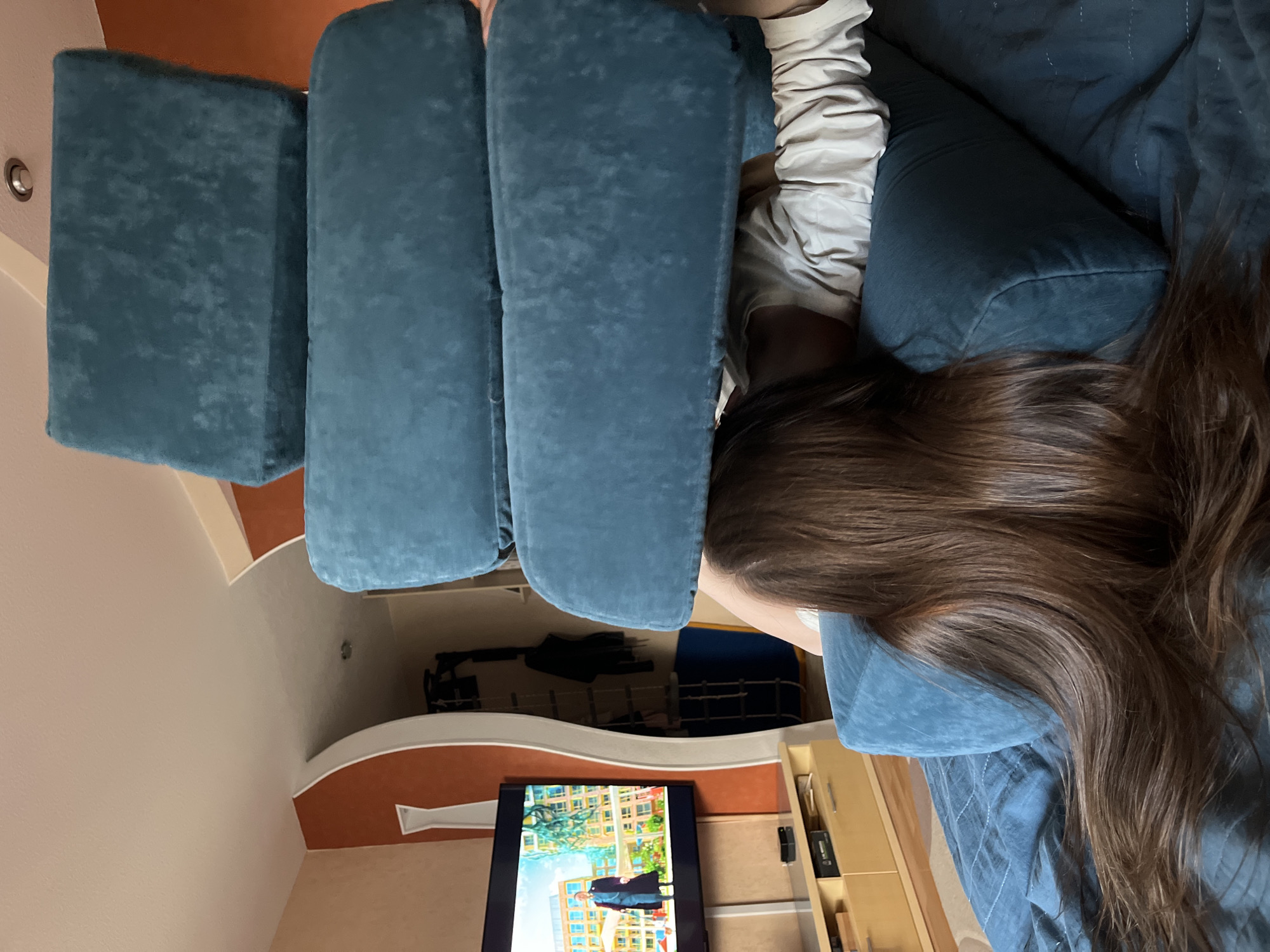 A person with long, brown hair is lying on a blue couch. The person is wearing a light-colored shirt. A television is visible in the background, displaying a cityscape. There is furniture against a wall in the background. The image was taken indoors. The location of Chaikovsky, Russia cannot be confirmed based on the image.