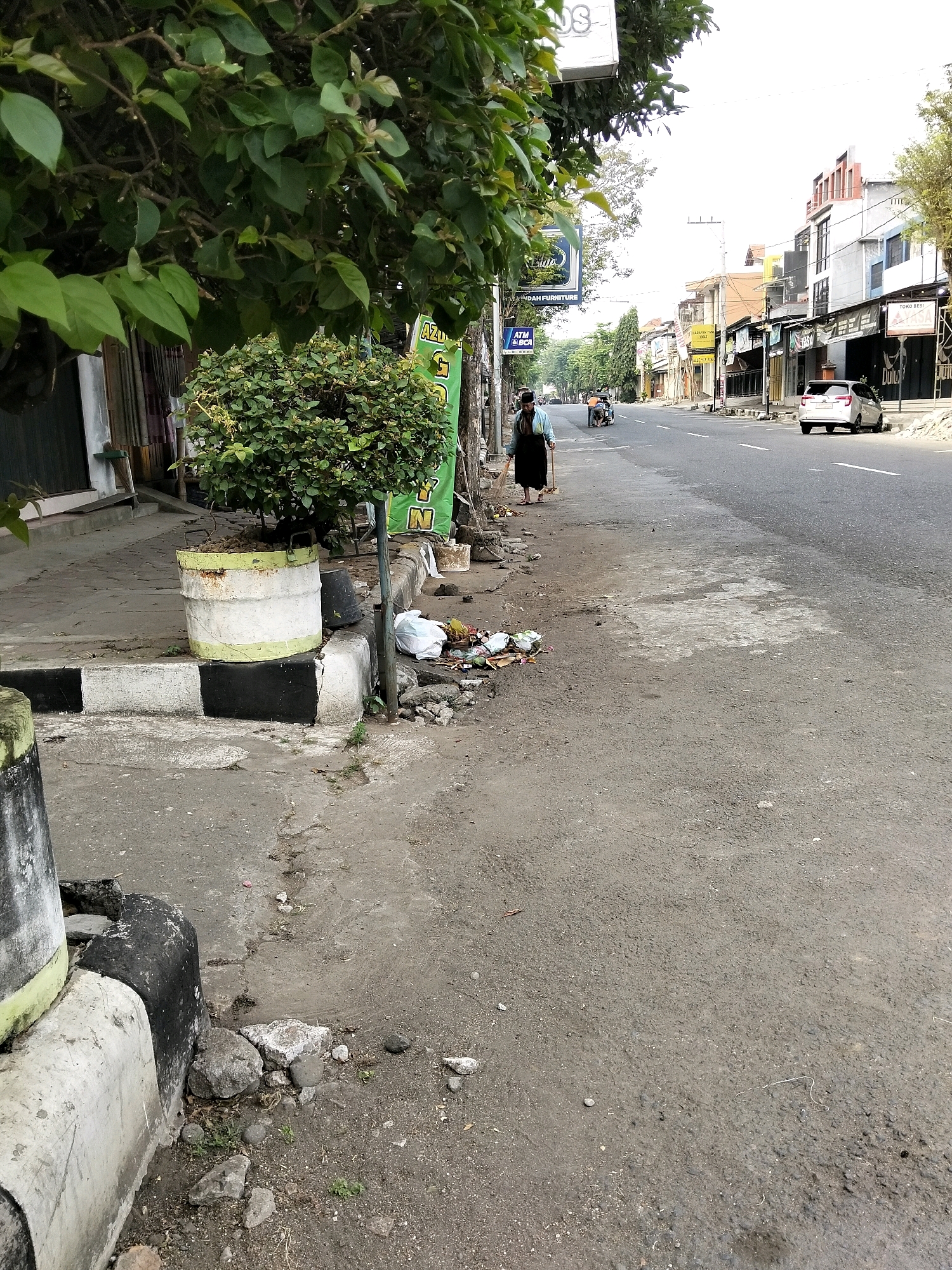 The image depicts a street scene in Purworejo, Indonesia, during daytime. The foreground shows a sidewalk with a large potted plant, a concrete curb painted black and white, and some scattered rocks and debris. A person dressed in a dark garment is seen sweeping the sidewalk with a broom. Further down the street, other individuals are visible, one on a motorcycle. The street itself is paved and appears to be a commercial area with buildings lining both sides. Some of the buildings have signs, indicating shops or businesses. A white car is parked on the right side of the street. The sky is overcast, casting a diffused light over the scene. The overall impression is that of a typical urban environment in Indonesia, with elements of daily life and commerce.