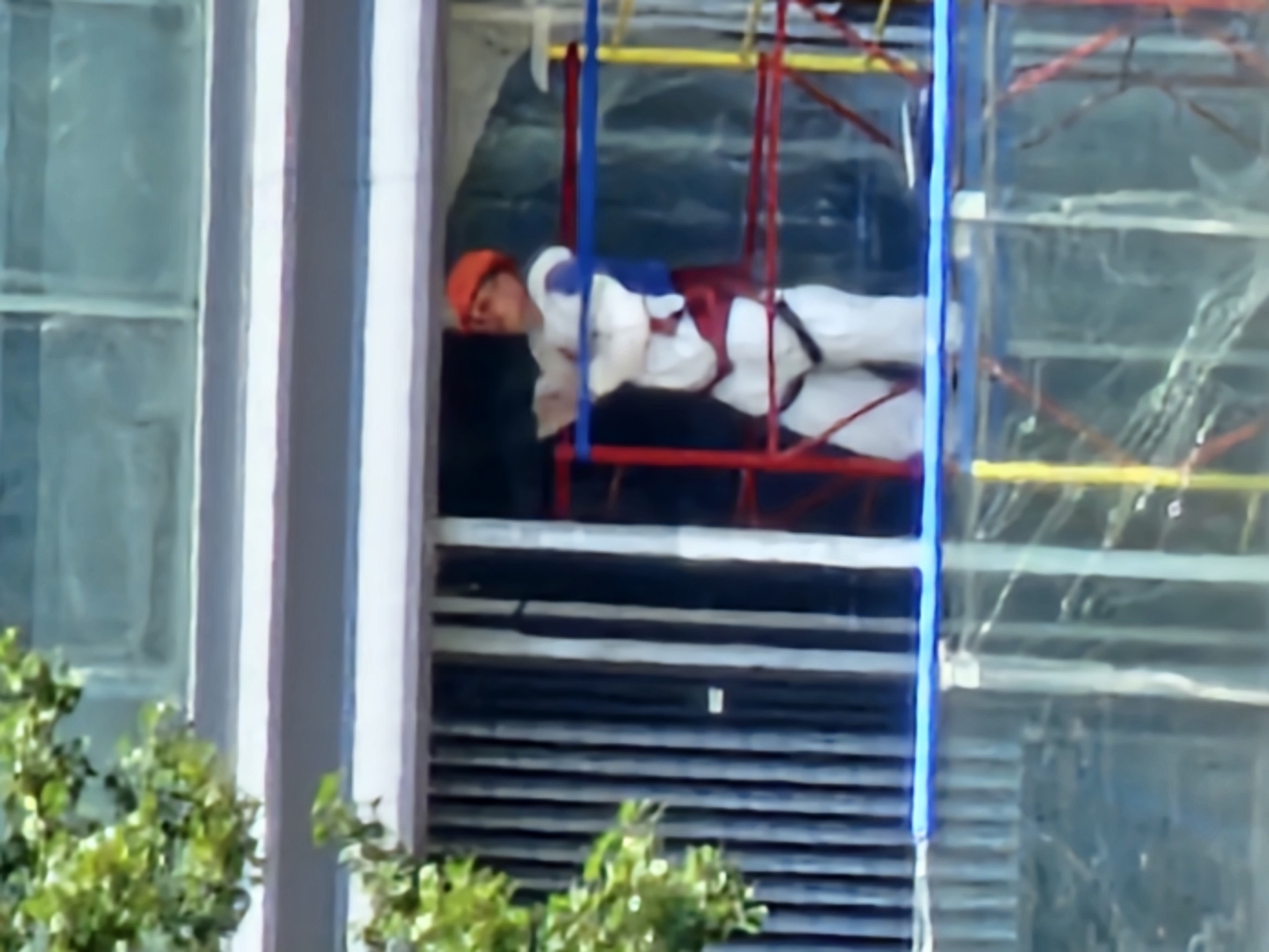 In Moscow, Russia, a person is seen on scaffolding outside a building. The individual is wearing a white protective suit, a blue shirt, and an orange helmet. They are positioned within a red, yellow, and blue scaffolding structure, appearing to be engaged in some form of construction or maintenance work. The building behind them features dark vertical blinds and large windows. Green foliage is visible to the left of the frame, suggesting the presence of trees or plants near the building. The scene appears to be captured during daylight.