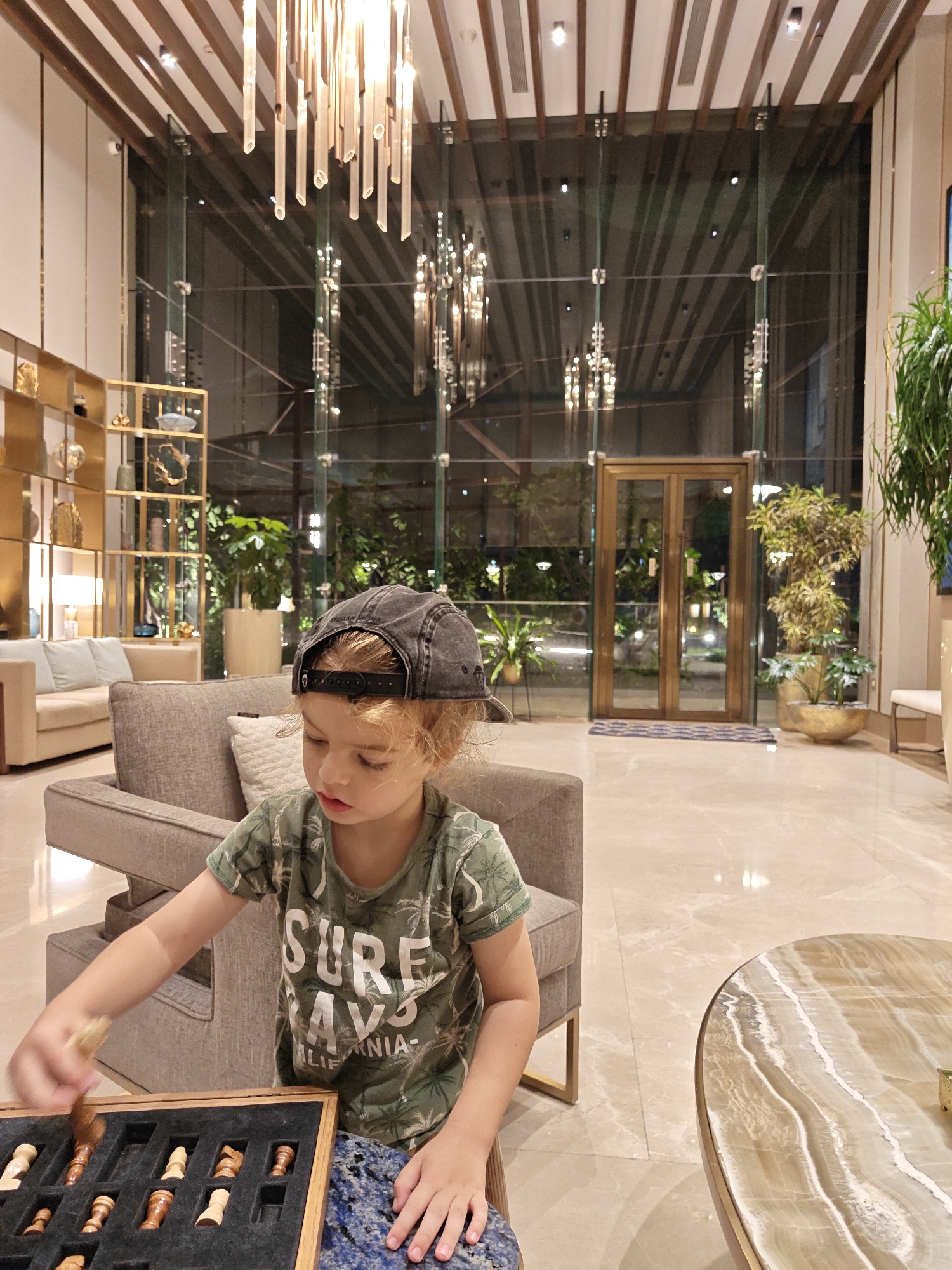 A young boy wearing a dark grey baseball cap and a green t-shirt is playing with chess pieces in a well-lit interior, presumably a hotel lobby. He is seated on a grey chair, placing a chess piece into a chess box. Next to him is a round marble table. The lobby features a high ceiling with decorative hanging light fixtures, large glass windows with plants visible outside, a light-colored tile floor, and decorative gold shelving. A beige couch is partially visible in the background. The photograph appears to have been taken in Mumbai, India.