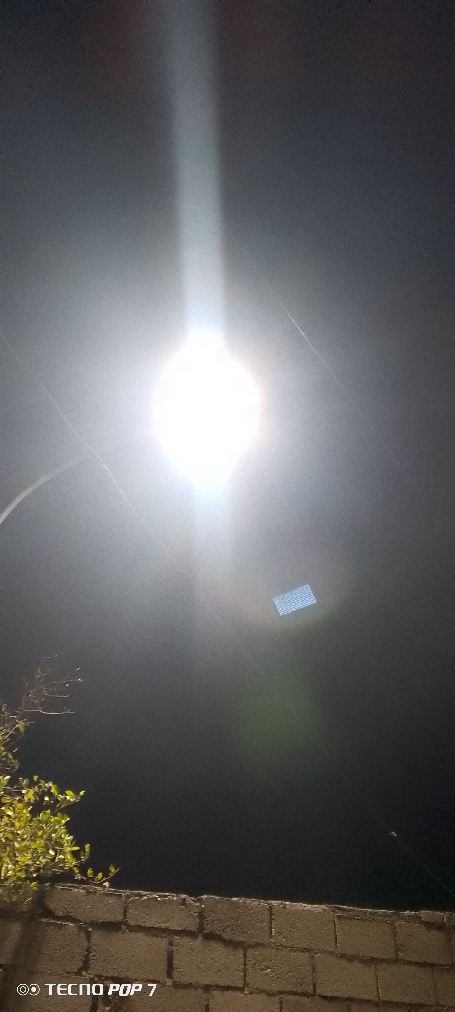 The image captures a night scene, presumably in Altagracia, Venezuela, based on context. A bright street lamp dominates the center of the frame, radiating intense light. A brick wall with foliage visible above sits at the bottom, suggesting an outdoor setting. Above the lamp, a rectangular object with a grid-like pattern is discernible, possibly a part of the light fixture. The camera used to capture the image is a Tecno Pop 7, evident from the watermark in the corner. The overall scene is dark, with the light source creating a strong contrast.