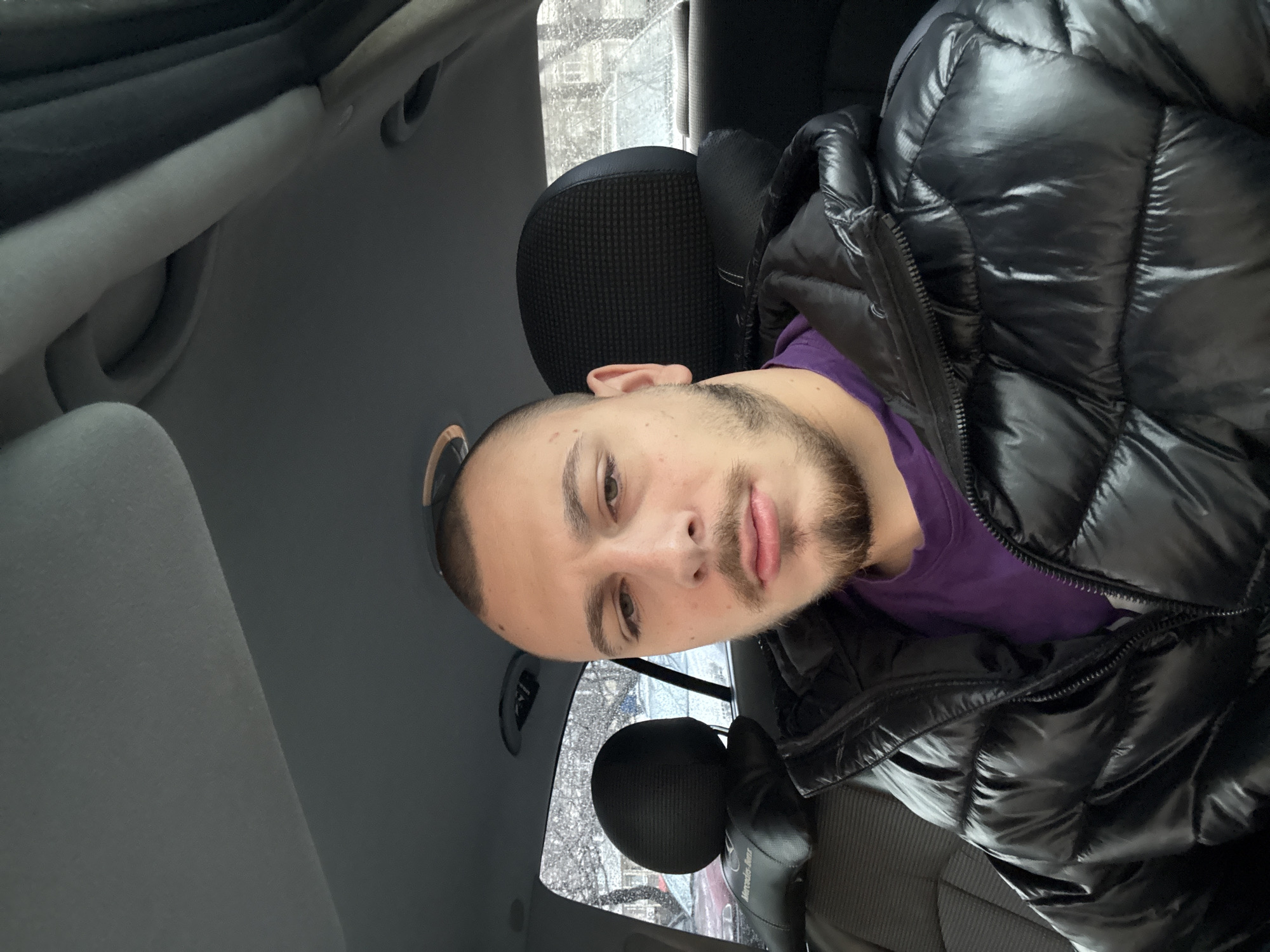 A man sits in the driver's seat of a car, wearing a shiny black puffer jacket over a purple shirt. The car interior is gray. The window shows a view of a snowy day in Stara Zagora, Bulgaria, with bare trees and buildings visible. The headrest behind the man is black and textured.