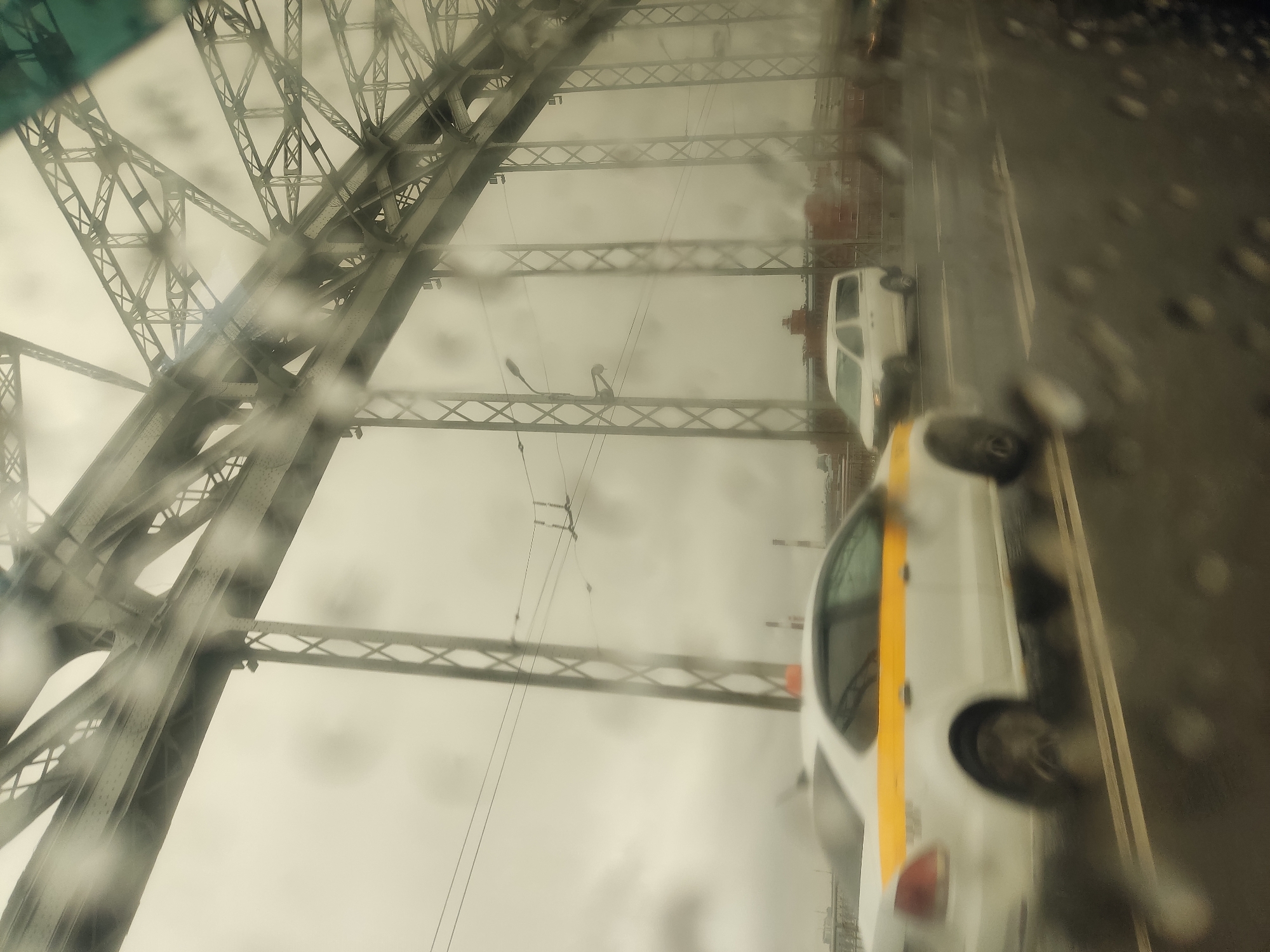 The image shows a view from a vehicle, possibly a car, on the Alexander Nevsky Bridge in Saint Petersburg, Russia, on a rainy day. The bridge's steel structure is visible on the left, obscured by raindrops on the vehicle's windshield. Cars, including a taxi with a yellow stripe, are seen driving on the bridge. The overall environment is overcast and wet.