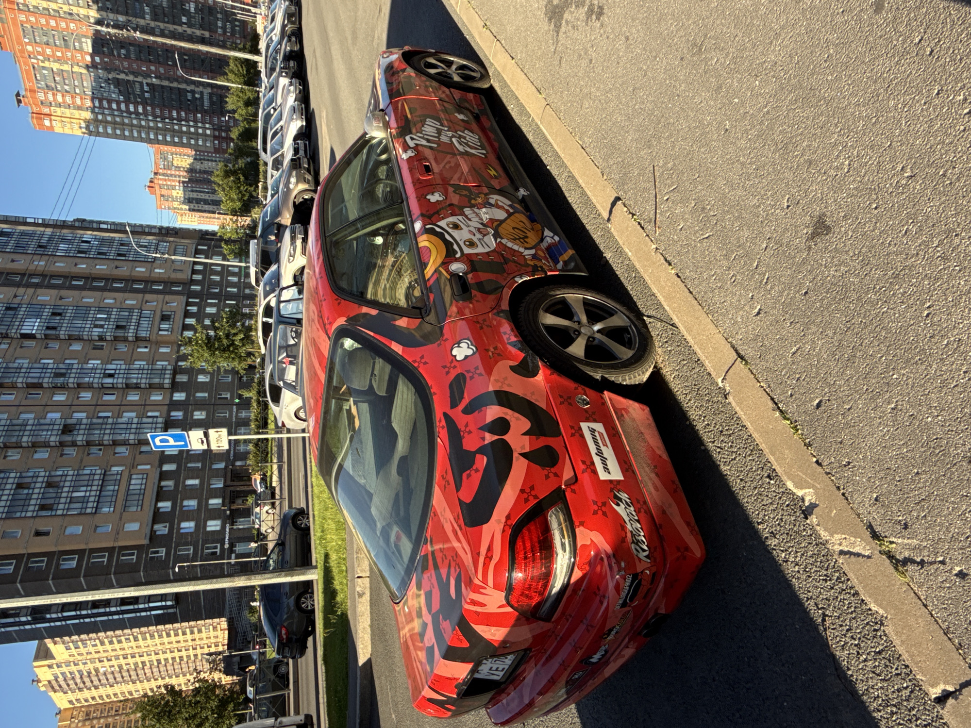 In Pargolovo, Russia, on a clear, sunny day, a modified red sedan with elaborate black and red graphic decals is parked on the right side of an asphalt street, facing away from the camera and to the left. The car features a cartoonish decal of a cat wearing a samurai hat and holding a baseball glove on its passenger side door, along with the text "Pimp my Ride." The rear of the car displays large, abstract black graphic designs. Adjacent to the car is a concrete curb and sidewalk, with patches of grass growing through cracks. In the background, large, multi-story residential buildings dominate the skyline, with a blue sky above. Other vehicles are parked and driving on the street in the distance. The overall scene depicts an urban environment with a focus on the uniquely decorated automobile.