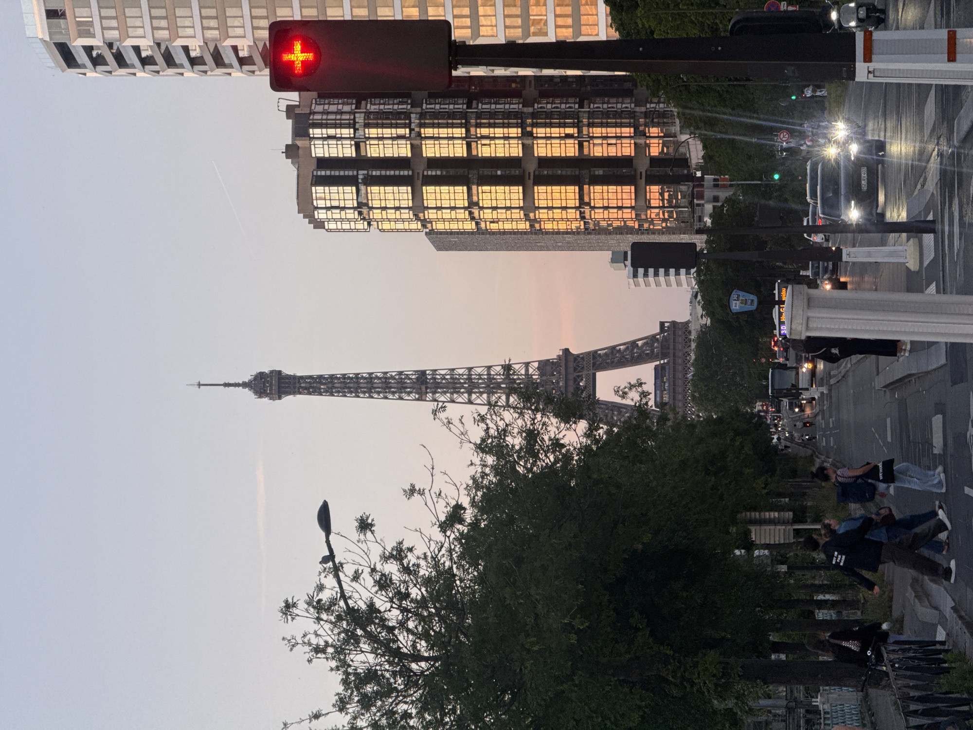 The image depicts the Eiffel Tower in Paris, France. The tower is seen in the distance against a backdrop of a pinkish sky, likely during sunset or sunrise. In the foreground, there is a tree with dark green foliage. To the right of the tree, people are seen walking on a sidewalk. A street with cars and traffic lights is also visible. The traffic light in the image is red. Buildings line the street, with a modern building reflecting the sunlight.