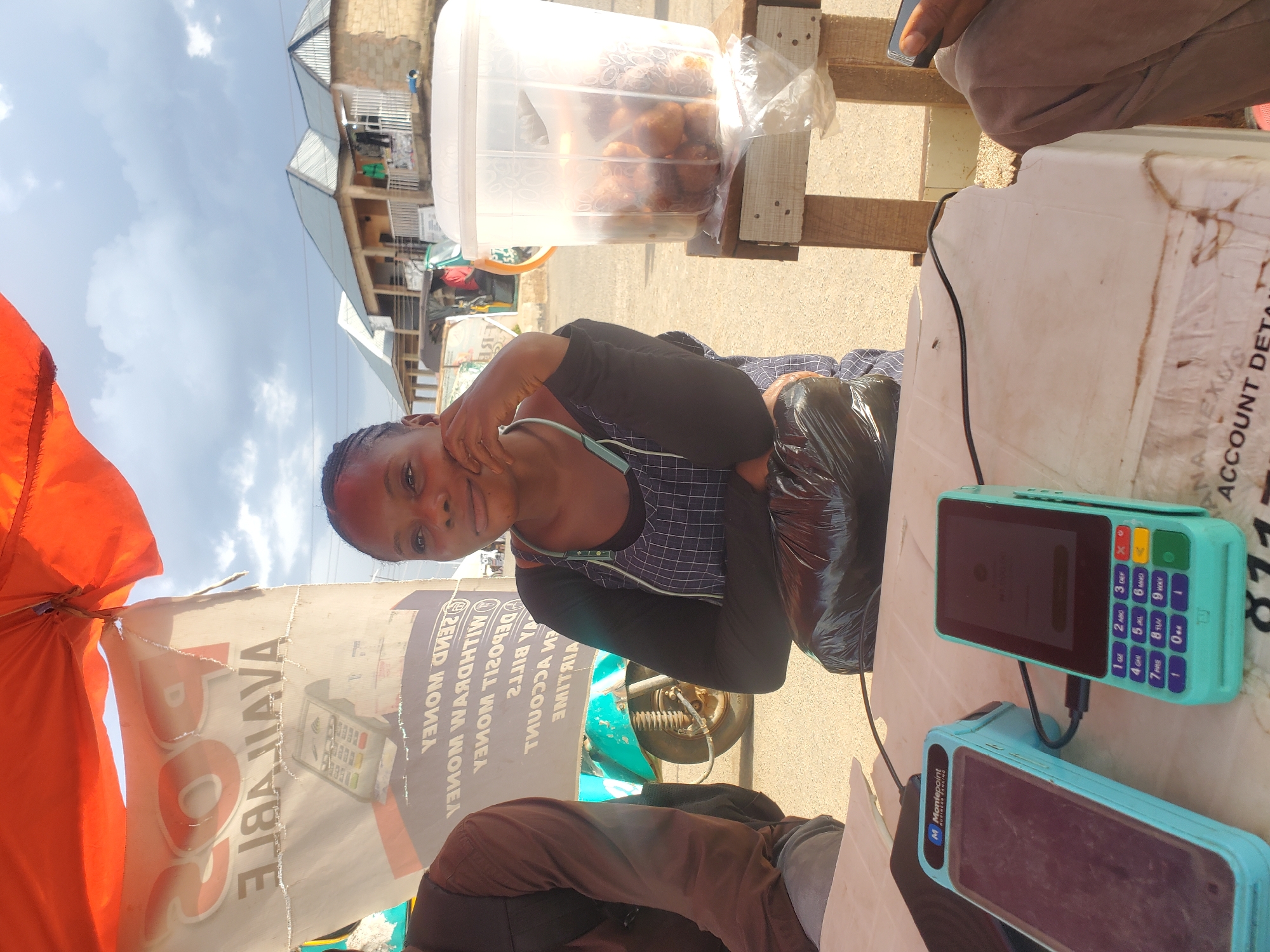 This media file shows a young woman at an outdoor market stall in Gwong, Nigeria. She is wearing a dark long-sleeved shirt with a plaid design across the chest and is holding a black plastic bag. The market stall appears to be selling food items, as a clear plastic container filled with fried snacks is visible. Two mobile payment terminals are also prominent on the counter, suggesting a transaction or business activity is taking place. One terminal is teal and displays a screen, while another, similar teal terminal is positioned below it. An orange umbrella is partially visible in the foreground. In the background, there are buildings with corrugated metal roofs and a bright sky. The lighting suggests it is daytime, and the overall scene depicts a busy, everyday market environment. Visible text includes "JAVA", "AVA", "READ WOWЕЛ", "MILHDBYM WOWЕЛ", "DELOILOVER", "M Moniepoint", "ANA NEXUS", and "ACCOUNT DET".