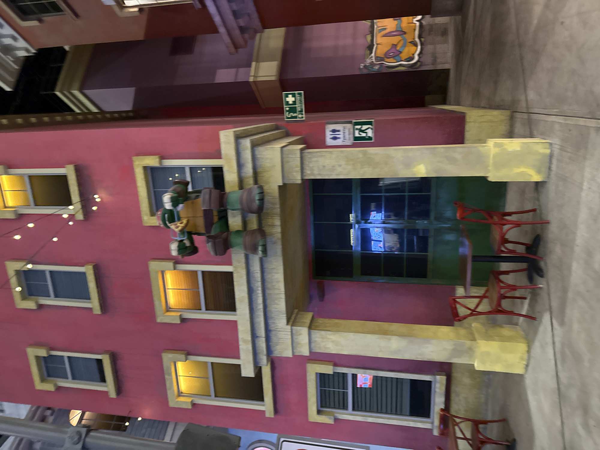The image presents an indoor commercial space, likely a food court or entertainment area in Moscow, Russia, designed to mimic an outdoor street scene. The central feature is a vibrant pinkish-red building facade, richly accented with yellow or gold trim around its numerous rectangular windows and architectural ledges. Several windows emit a warm, yellow light, suggesting an evening ambiance or controlled indoor lighting.
Decorative elements on the building include a string of small white lights, and a vertical stack of what appear to be barrels or planters, accompanied by a novelty pizza slice decoration. In front of the building, a dining area is set up with several red chairs clustered around small, dark tables, though no people are present. The floor surface is a plain, light grey, resembling concrete paving.
Various signs are affixed to the building: a green sign indicating "FIRST AID" with a white cross and an 'i' symbol, alongside a wheelchair accessibility symbol; a white sign for men's and women's restrooms; and a green "EXIT" sign with a running figure. In the background, above the main building, a colorful, graffiti-style mural is visible on a grey wall. The scene evokes a lively, urban setting, despite being contained within an indoor structure.