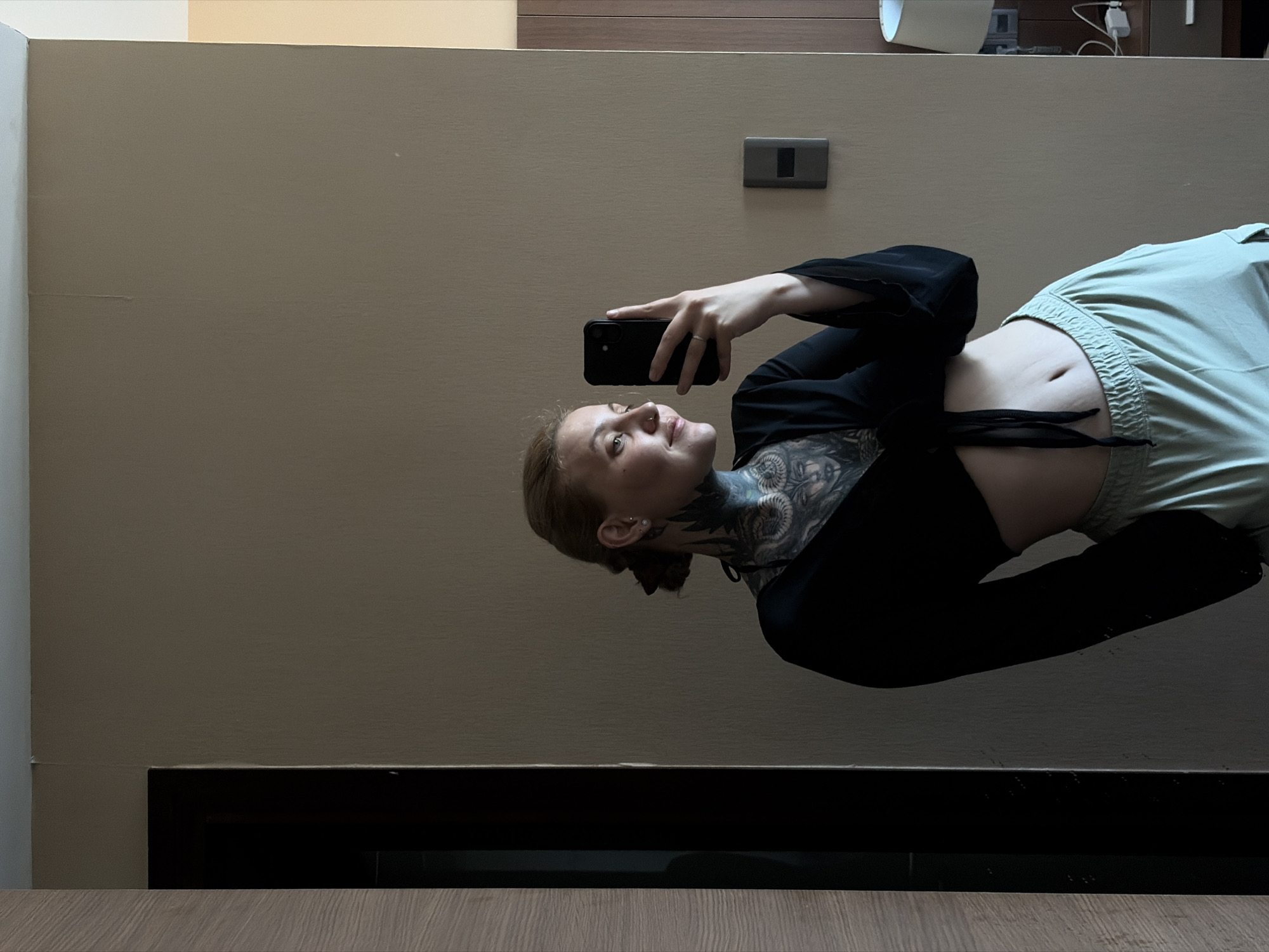 A woman is taking a selfie with her smartphone in a room. She is wearing a black crop top and light green sweatpants. Her neck is tattooed. The room has beige walls and a dark baseboard. A light switch is on the wall. The location is likely in Đà Nẵng, Vietnam, but this cannot be confirmed from the image alone.
