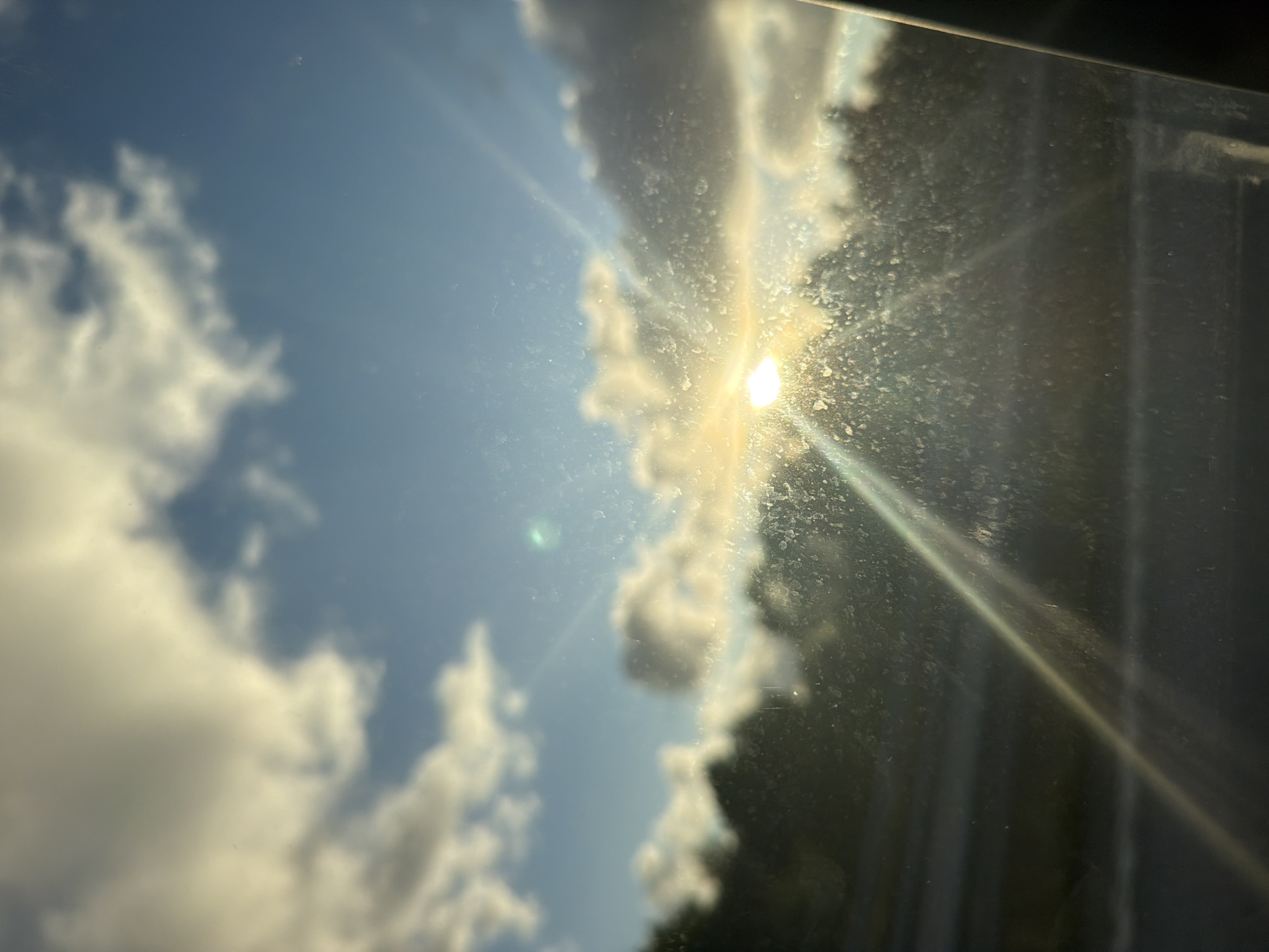 The image depicts a sunlit scene viewed through a dirty window. The sun is positioned in the upper center of the frame, casting bright rays that streak across the image. Clouds, both bright and dark, are visible in the sky, suggesting a partly cloudy day. Below the sky, a silhouette of trees lines the horizon. The foreground is dominated by the window, which is covered in numerous smudges and streaks, obscuring a clear view of the landscape. The overall impression is one of a bright, possibly early morning or late afternoon, sky, seen from inside a vehicle or building. The location is Ödsmål, Sweden.
