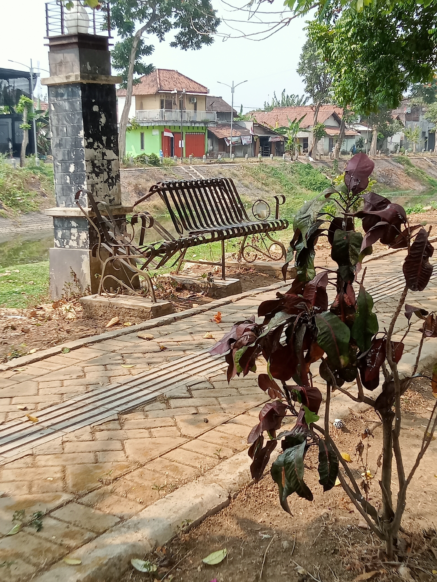 The image depicts an outdoor scene in the city of Tulungagung, Indonesia. In the foreground, a stone pathway is visible, with a textured, raised strip running parallel to it, suggesting it is a tactile paving for the visually impaired. Adjacent to the pathway is a patch of dry soil with scattered fallen leaves. A plant with dark red and green leaves stands to the right of the pathway.
Further into the image, a black metal park bench with decorative scrollwork is placed on a raised concrete base. Behind the bench, a decorative stone pillar with peeling black paint is visible. In the background, a body of water, possibly a canal or small river, is seen with grassy banks. Beyond the water, a row of residential buildings with terracotta-tiled roofs and muted-colored walls are present. Trees with green foliage are scattered throughout the scene, and the sky is overcast. The overall impression is one of a quiet, possibly urban park area.