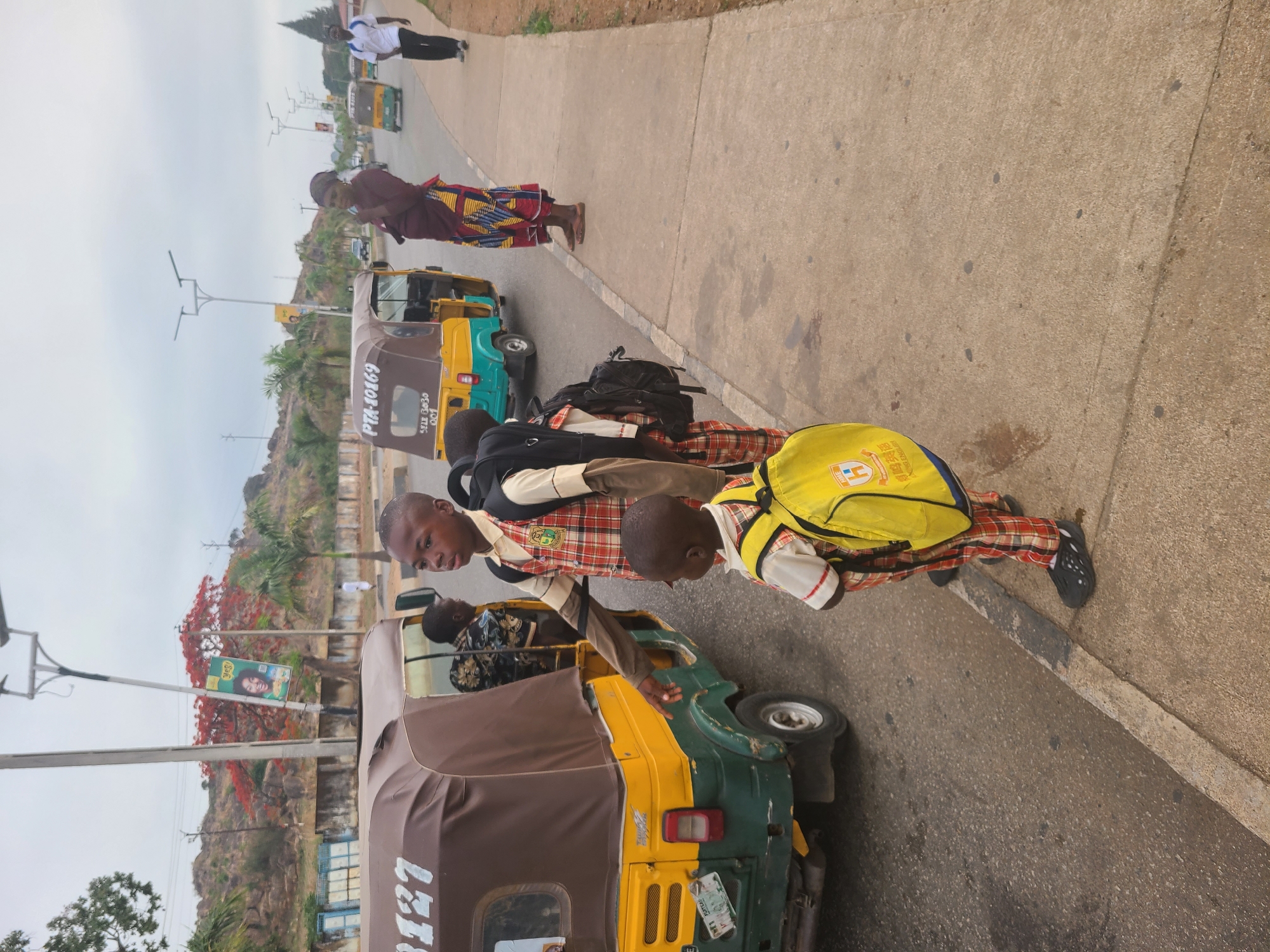The image captures a street scene in Jos, Nigeria, with a focus on two schoolboys dressed in plaid uniforms and backpacks, appearing to interact with a local "keke" or auto-rickshaw. One boy, with a yellow backpack, leans into the vehicle, while the other, wearing a black backpack, stands beside him. Several other auto-rickshaws are visible along the street. A woman in traditional African clothing stands on the sidewalk nearby. In the background, a rocky hill, trees, and a billboard are visible. The sky is overcast.