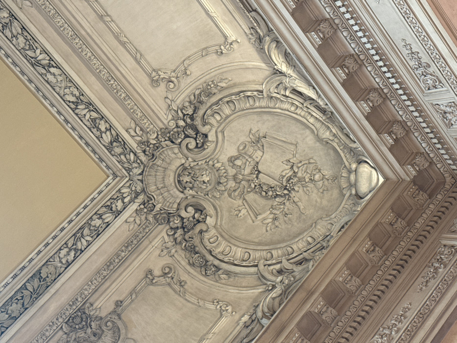 This image captures an upward-angled view of a highly ornate ceiling and upper wall section within the El Ateneo Grand Splendid bookstore, located in Buenos Aires, Argentina. No individuals are present in the scene.
The central focus is a large, light beige, oval-shaped plaster relief on the ceiling, depicting two open books surrounded by a detailed floral garland with leaves and small fruits. Behind these, a rolled scroll or parchment secured by a ribbon is visible. This central motif is framed by elaborate, undulating rococo-style scrollwork, acanthus leaves, and additional floral elements, including a shell-like motif at the top and a stylized vase at the bottom.
Surrounding this central relief, the ceiling features multiple layers of moldings. An inner band displays a repeating geometric pattern, followed by a flat panel. Towards the corner visible in the upper sections of the image, the ceiling transitions to a plain, lighter beige surface, bordered by a wide, dark molding intricately carved with a continuous pattern of stylized leaves and branches.
Below the ceiling, a substantial cornice connects to the upper wall. This cornice comprises several horizontal bands of molding, including one prominent section with regularly spaced small decorative brackets or corbels alternating with recessed rectangular sections. Further down, additional moldings exhibit repeating patterns such as small circles and stylized leaves. The upper wall section below the cornice appears in a slightly warmer, light reddish-brown or terracotta hue, contrasting with the lighter tones of the ceiling. The entire surface demonstrates intricate three-dimensional plasterwork under soft, diffused lighting.