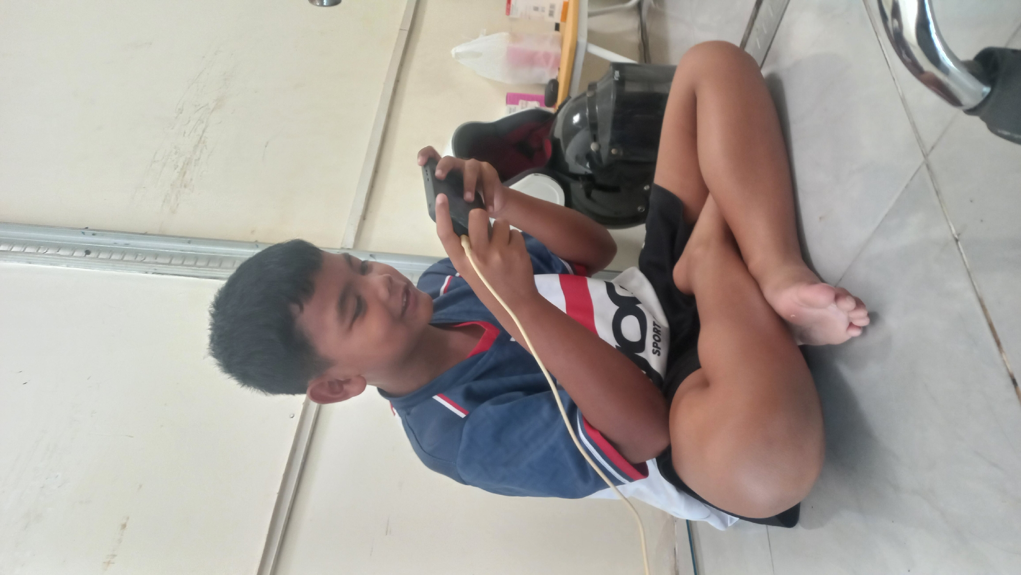 A young Indonesian boy is sitting cross-legged on a tiled floor, playing a video game on a handheld console. He is wearing a blue and white t-shirt with red trim and black shorts. His left leg is bent and his foot is resting on the floor, while his right leg is crossed over his left thigh. A white cable is plugged into the console and runs out of frame to the left. In the background, a black helmet is propped up on a shelf or stand, and a small table with some items on it is visible. The scene appears to be indoors, possibly in a home or a common area. The environment is relatively simple and unfurnished. The boy is focused on his game and smiling. The overall impression is one of casual leisure. The location is identified as Jetis Indah, Indonesia.