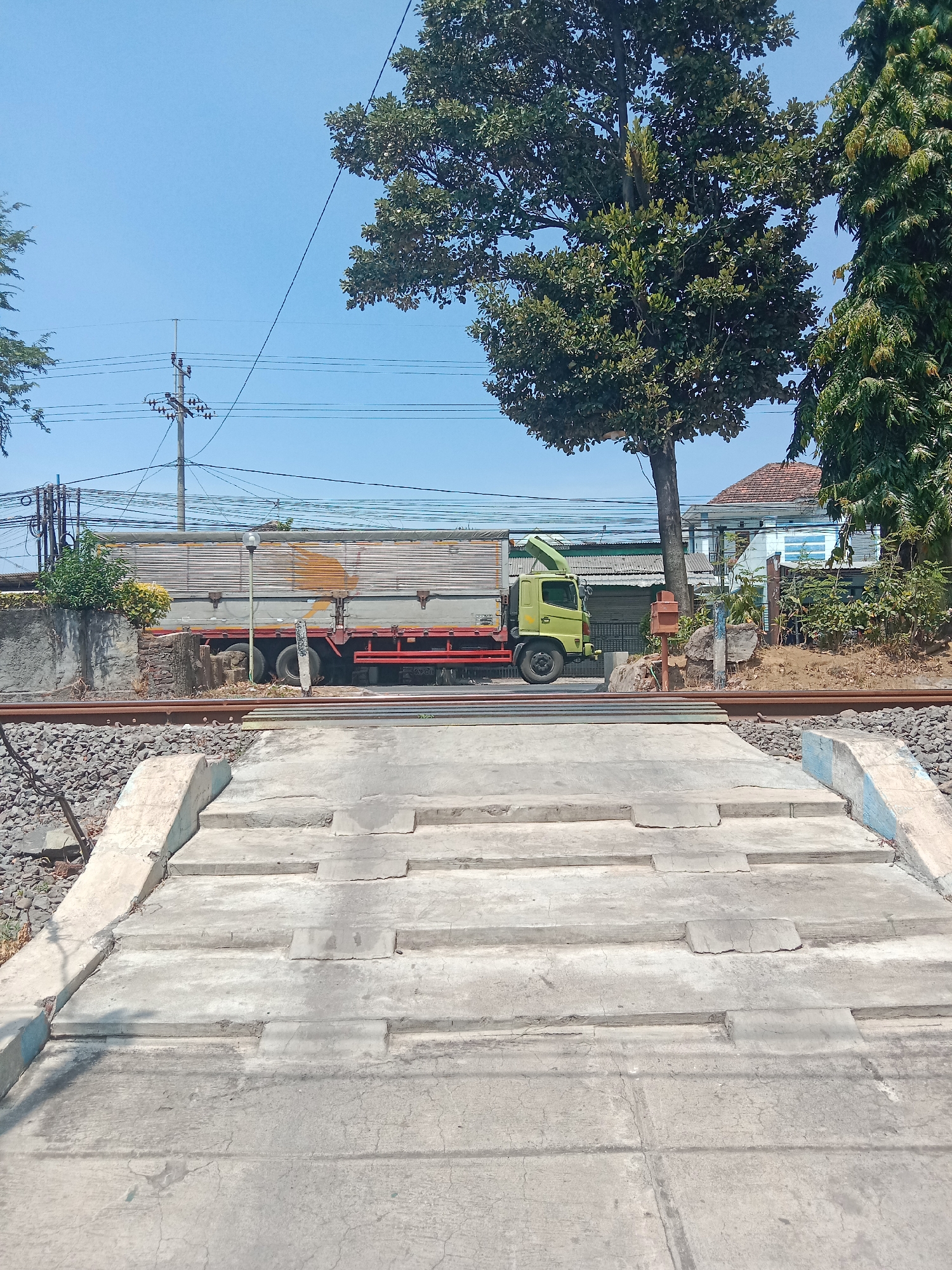 The image shows a railway crossing in Probolinggo, Indonesia. In the foreground, there is a concrete pedestrian crossing with several steps. Beyond the crossing, railway tracks run horizontally. A light green truck with a silver cargo bed is stopped at the crossing. Behind the truck, there are some buildings, power lines, and trees. The sky is clear and blue.