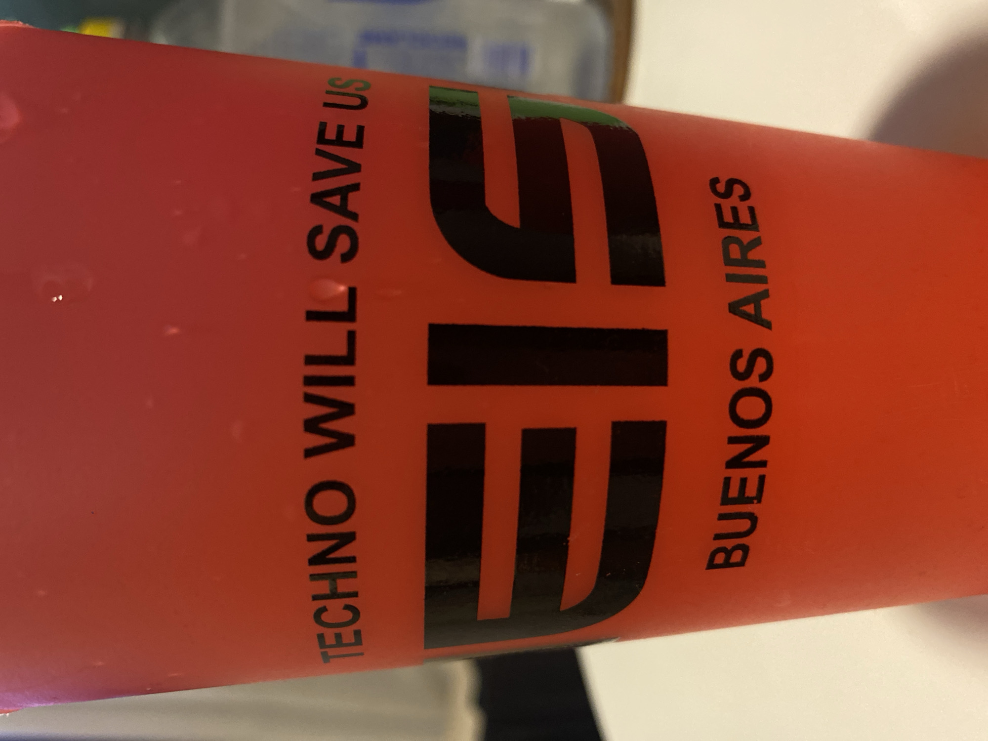 The image depicts an orange cup with black lettering that reads "TECHNO WILL SAVE US" vertically on the left side. Next to it, the black letters EIF are printed vertically. To the right of that, "BUENOS AIRES" is also printed vertically. The background shows blurred objects, possibly a bottle or container. The overall scene appears to be indoors.