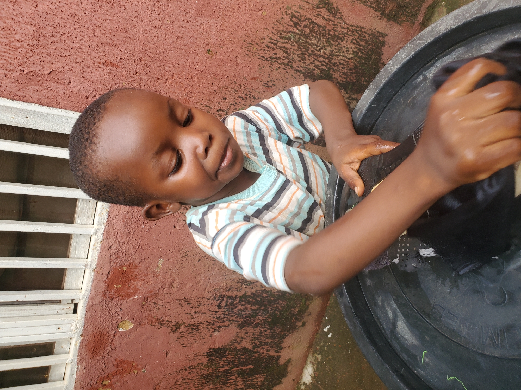 A young Black child with short hair, wearing a horizontally striped t-shirt in blue, white, orange, and black, is captured in a close-up, high-angle shot. The child is looking down with eyes closed, and their mouth is slightly open, suggesting concentration or exertion. They are interacting with a large black basin filled with water. The child's hands are wet and appear to be washing a piece of black fabric.
The setting is outdoors, evident from the textured, reddish-brown wall in the background, which shows signs of weathering and some dark patches that could be moss or dirt. To the left of the child, a white slatted window grate is visible, suggesting the proximity of a building. The ground around the basin is wet and appears to be made of concrete or similar material.
The lighting suggests it is daytime, with no strong shadows indicating a specific time of day. The overall scene evokes a sense of everyday activity, possibly a chore or playtime involving water. The child's expression and engagement with the task are the focal points of the image. No visible text is present in the image. The location context provided is Jos, Nigeria.