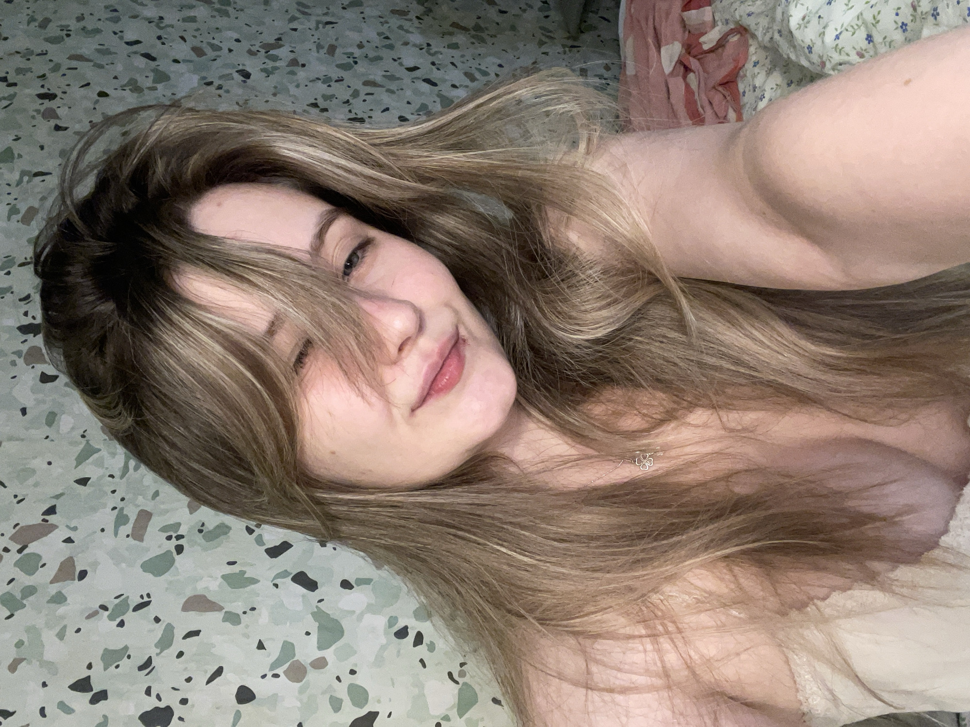 A young woman with long, blonde hair covering part of her face, is looking at the camera with a slight smile and one eye closed. She is wearing a cream-colored, lace-trimmed top and a delicate necklace. The background is a wall with a terrazzo-like pattern in shades of green, beige, and black. She is positioned in what appears to be a bedroom, with bedding visible in the lower right corner of the frame.