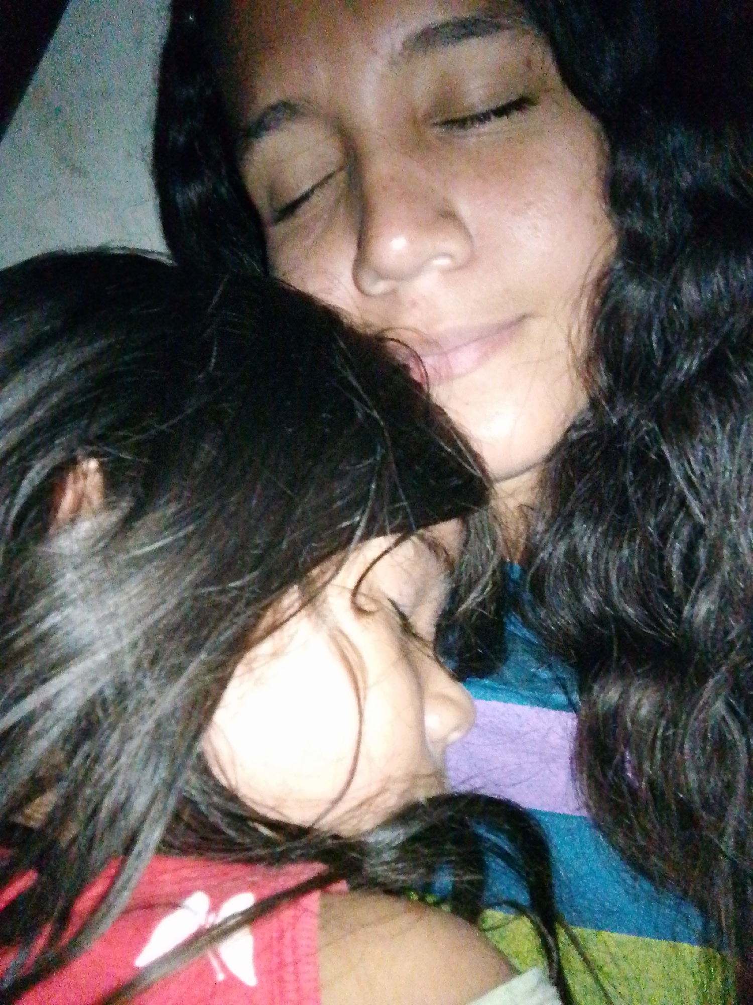 The image depicts two individuals, likely an adult female and a child, asleep or resting in close proximity, suggesting an intimate, indoor setting in Juan Griego, Venezuela. The upper individual, appearing to be an adult female, has dark, wavy hair and a medium complexion, with her eyes closed. She is wearing a garment with horizontal stripes in shades of blue, purple, and green. Her head is angled slightly upwards and to the left. The lower individual, a child, has dark, straight hair and is positioned with her head resting against the upper individual's shoulder/chest area, largely obscured by her own hair. Her eyes are also closed. She is wearing a red garment featuring a white graphic element, possibly wings, on the front. The lighting is dim, contributing to a soft-focus effect across the scene.