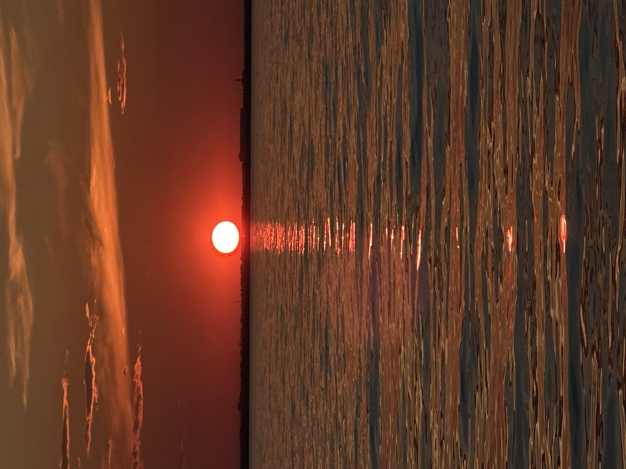This media file captures a serene sunset over a body of water, likely a lake or ocean, given the presence of rippling waves reflecting the sun's warm glow. The image is oriented vertically.
The left side of the frame features the sky, dominated by a vibrant orange hue. Wispy clouds, illuminated from behind by the setting sun, streak across the upper portion, creating a dramatic contrast with the deeper orange of the sky. The sun itself is a prominent, bright red-orange orb, positioned near the center of the left half, casting its light downwards.
The right side of the frame is dedicated to the water's surface. The waves are rendered as vertical lines of varying shades of orange, brown, and dark blue, creating a textured, almost abstract pattern. A bright, shimmering reflection of the sun stretches across the water from the sun's position in the sky, appearing as a luminous pathway. The light on the water suggests it is relatively calm, with small, consistent ripples.
The overall mood of the image is peaceful and tranquil. The rich colors of the sunset evoke a sense of warmth and natural beauty. The time of day is clearly sunset, characterized by the low angle of the sun and its intense, warm colors. No people, objects, or text are visible in the frame, focusing entirely on the natural elements of the sky and water. The location context of Kokshetau, Kazakhstan, suggests this could be a sunset over one of its many lakes or potentially the Ishim River. The weather appears clear, allowing for an unobstructed view of the sunset and its reflections.