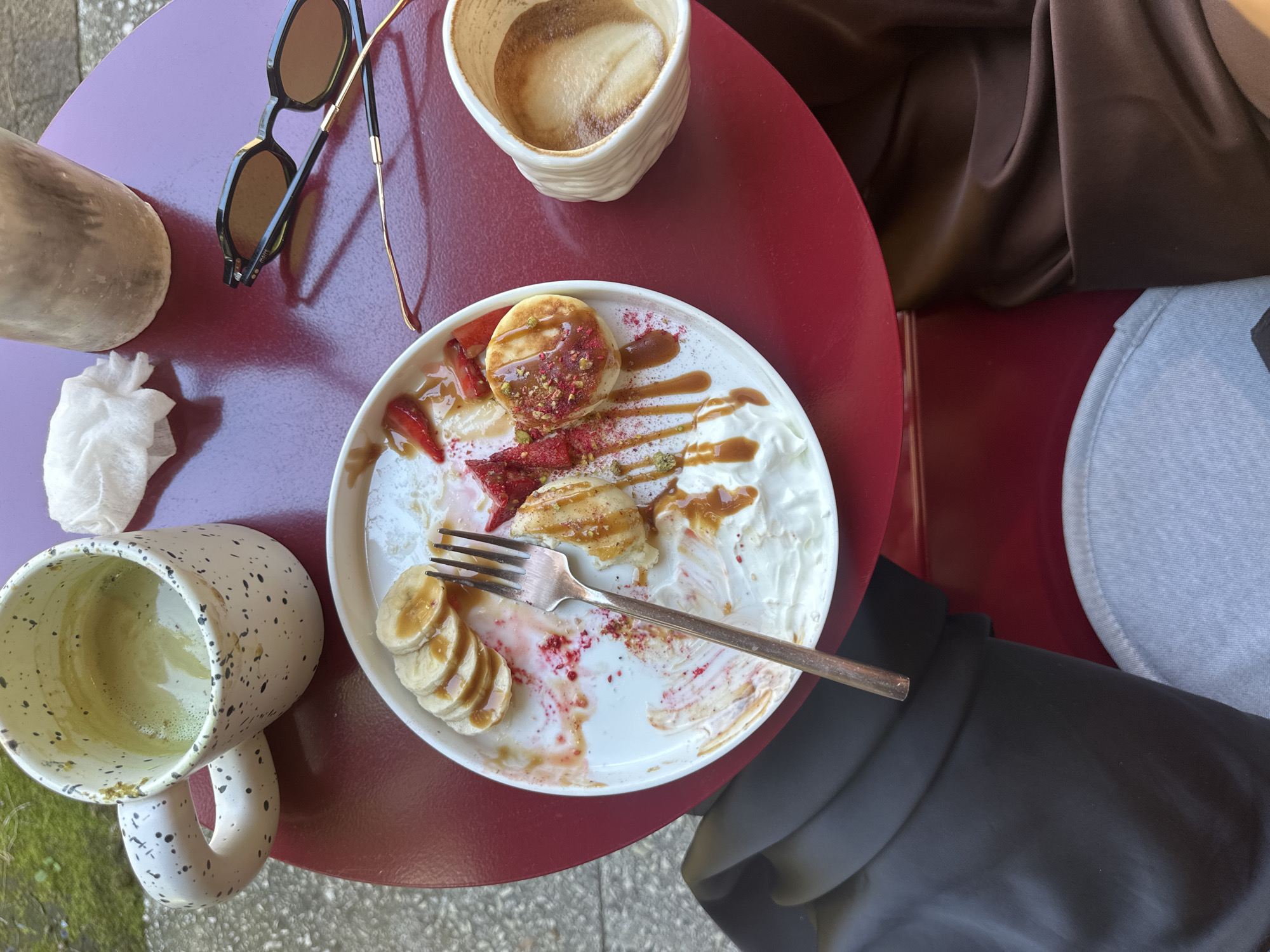 The image captures a close-up, overhead view of a red, round outdoor table at a cafe, with a portion of two people seated at it. On the table, a white plate holds partially eaten pancakes, sliced bananas, strawberries, whipped cream, a drizzle of caramel sauce, and sprinkled pistachios and red flakes. A fork rests on the plate. To the left, a white mug with black speckles contains a green beverage, likely matcha. To the right, a small white cup holds a coffee with latte art. A crumpled white napkin and a pair of sunglasses are also on the table. The background includes hints of outdoor greenery and pavement. The people are represented by their legs and lower torsos, clad in dark pants and a brown garment, suggesting a casual setting. No specific Sochi landmarks are visible.