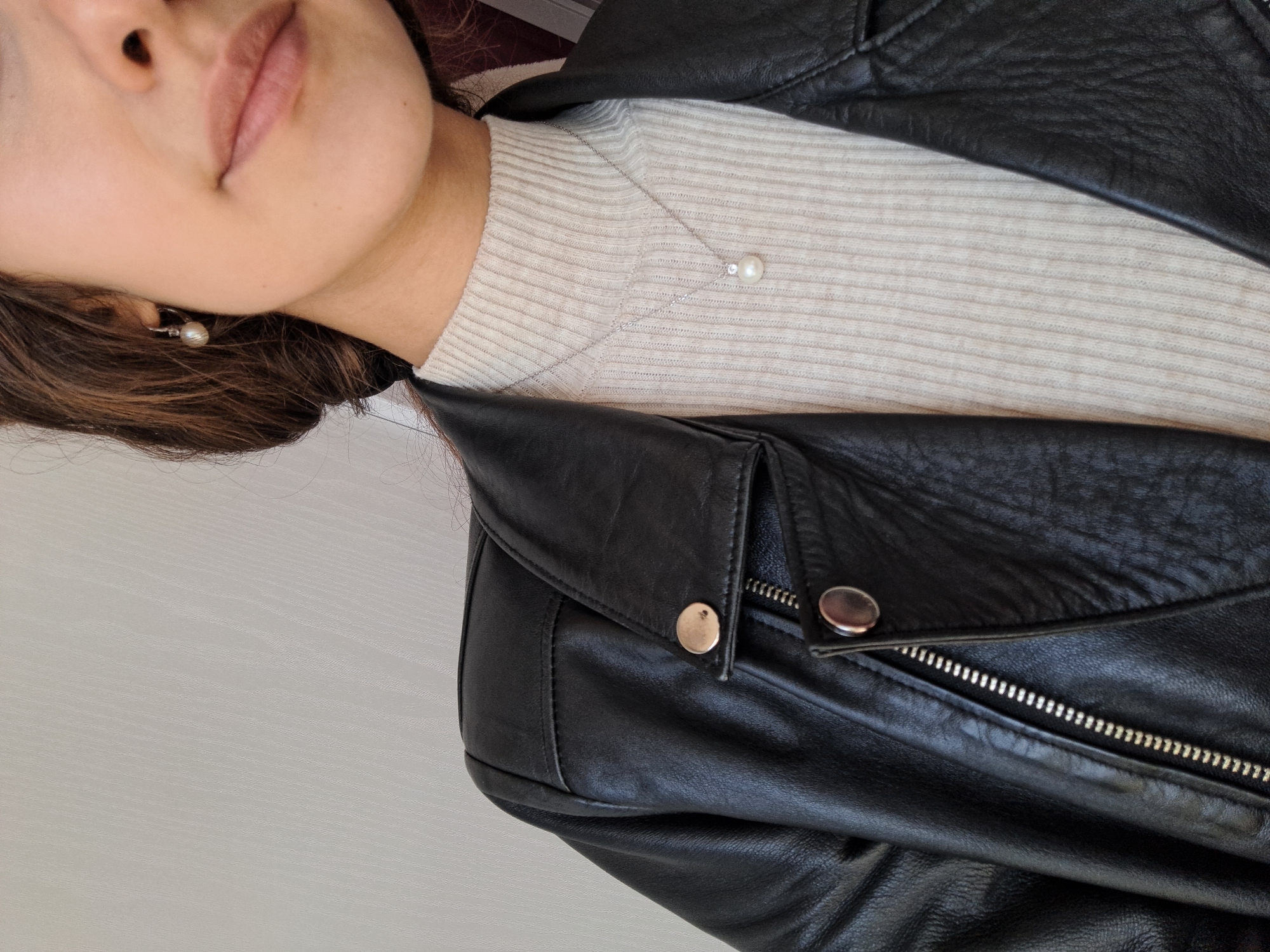 The image shows a person wearing a black leather jacket over a ribbed cream-colored sweater. The person has fair skin and brown hair, and is wearing pearl earrings and a pearl necklace. The setting is indoors.