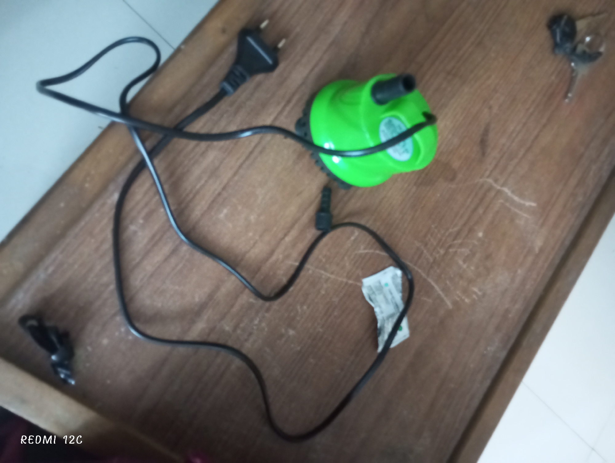 The image shows a wooden table surface with a green water pump and its black power cord arranged on it. The power cord includes a European plug. Also present are a set of keys and a small black clip. The photograph was taken by a Redmi 12C. The location is Lhokseumawe, Indonesia.