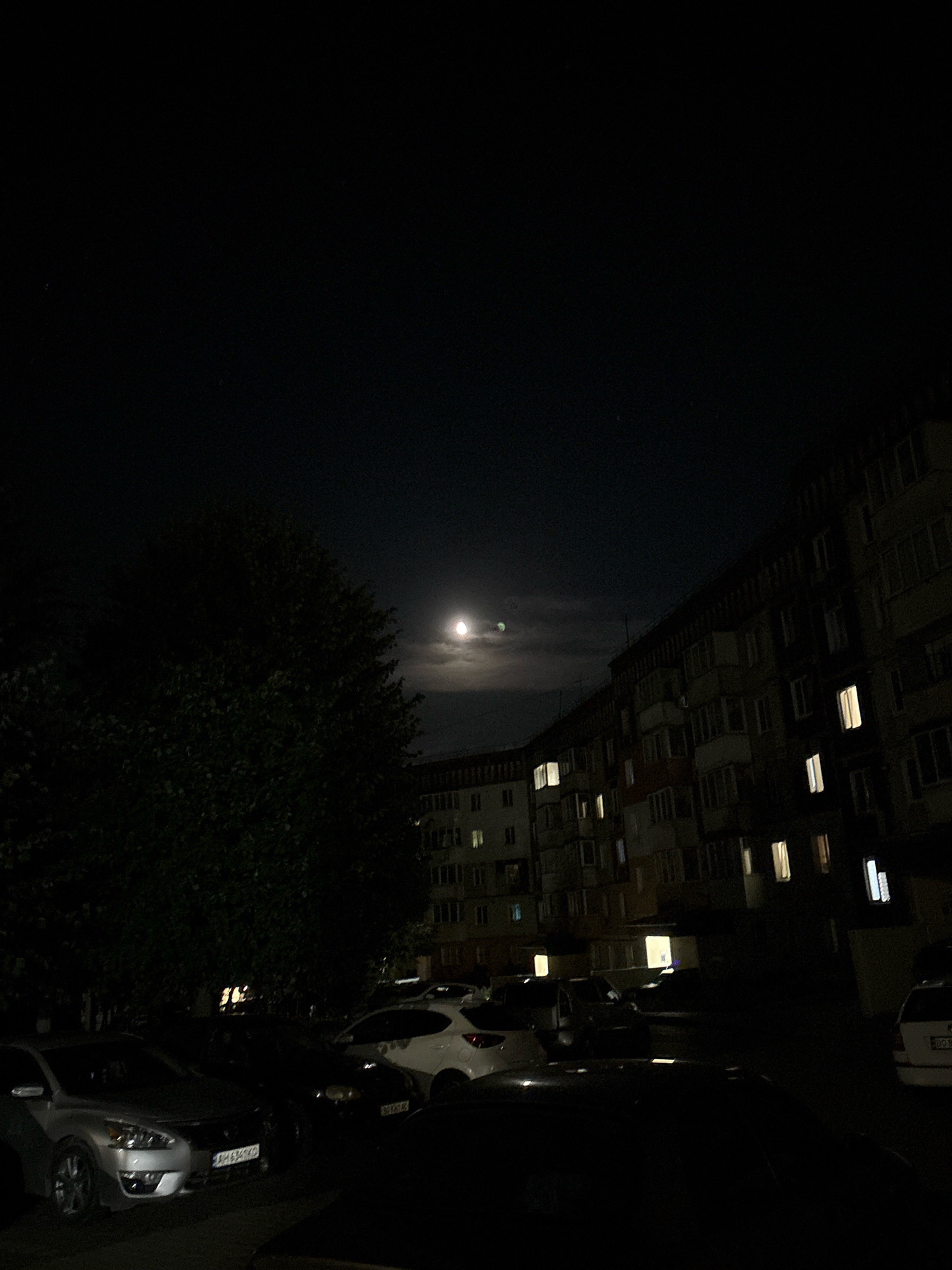 The image depicts a nighttime scene in an urban environment, likely a residential area in Ternopil, Ukraine. A large, dark tree dominates the left side of the frame. In the background, a multi-story apartment building with numerous illuminated windows is visible. The sky is dark, with a partially obscured moon visible in the upper center, surrounded by faint clouds. In the foreground, several cars are parked. The overall atmosphere is dark and quiet, illuminated by the moon and the lights from the apartment building.
