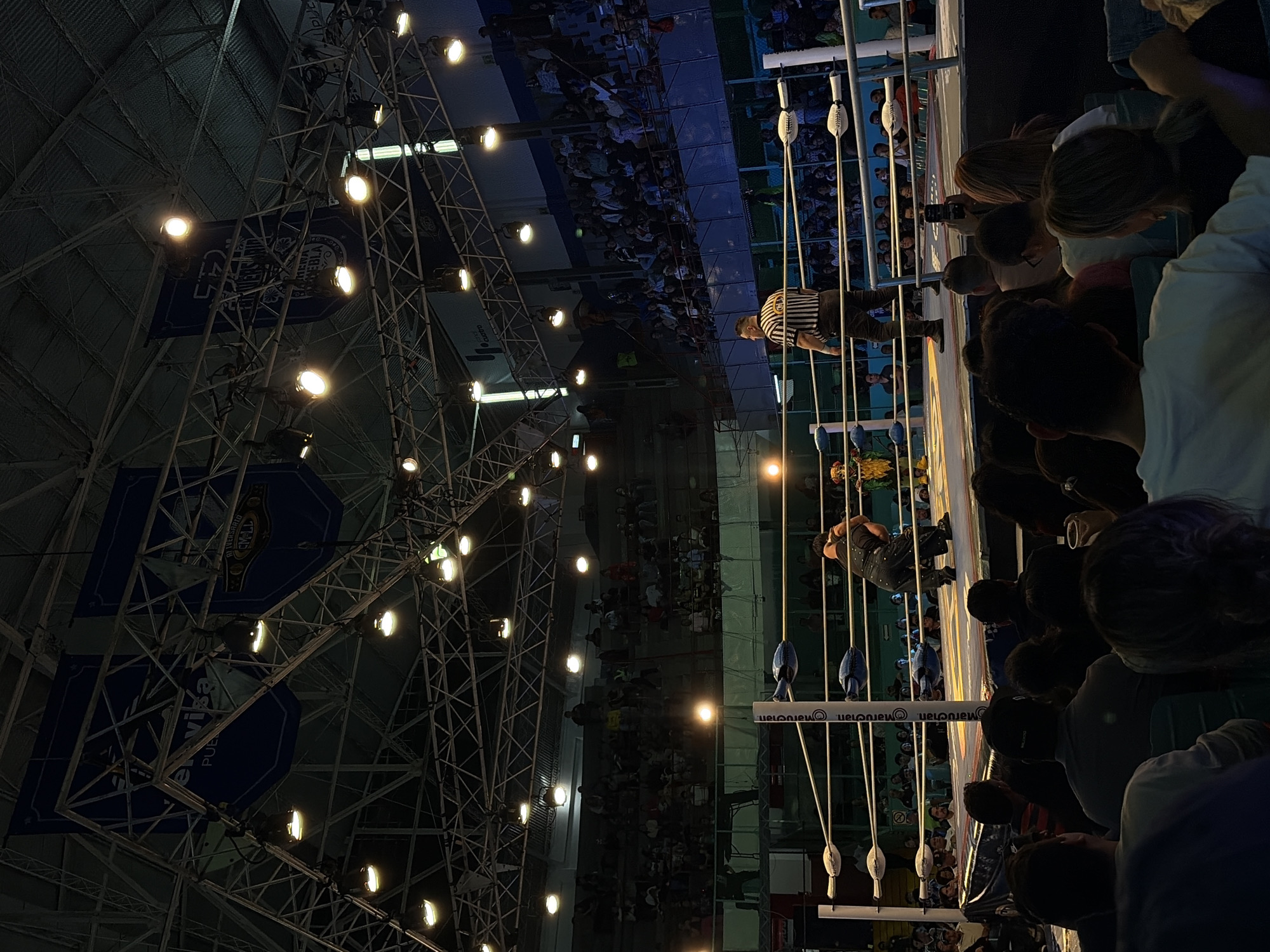 The image depicts an indoor wrestling event at the Gimnasio Miguel Hidalgo in Puebla, Mexico. In the foreground, a crowd of spectators is visible, many facing away from the camera, watching the event. The wrestling ring is the central focus of the middle ground, with two wrestlers engaged in a match. One wrestler, dressed in black overalls, is on his knees, appearing to be pinned or held down. The other wrestler, wearing a yellow and green outfit, stands over him. A referee in a black and white striped shirt stands to the side of the ring, observing the match. The background shows tiered seating filled with more spectators and the gymnasium's overhead lighting and structural elements, including banners hanging from the ceiling. The overall atmosphere is one of an active sporting event.