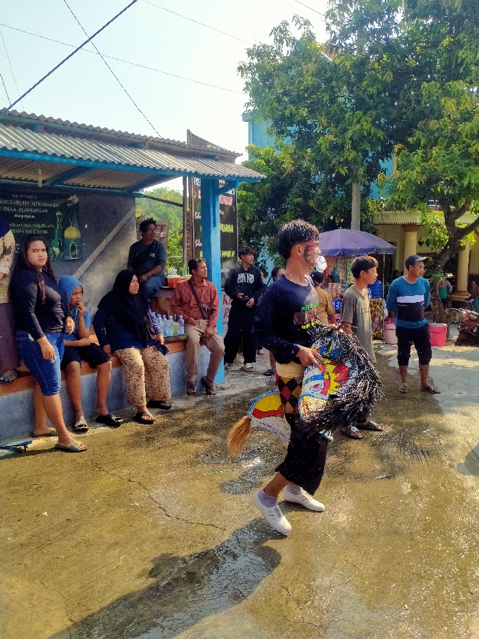 The image captures an outdoor scene in Mekarwangi Village, Blimbingan Village, within Sepat, Indonesia, under bright daylight. A young male, characterized by face paint on his cheeks and a dark, curly hairstyle, is depicted in motion, wearing a dark long-sleeved shirt, black pants, and white shoes. He holds a colorful, stylized horse prop, consistent with a Jaran Kepang or Kuda Lumping performance item, featuring a dark, shaggy mane and a reddish-brown tail-like attachment. The ground is a wet, cracked concrete surface reflecting sunlight. To the left, under a shelter with a corrugated metal roof and blue supports, multiple individuals are seated on a concrete ledge, observing the scene. This group includes a female in a black top and blue jeans, a male in a blue hooded sweatshirt, two females wearing headscarves, and two adult males. Plastic water bottles are visible near the seated individuals. In the background, large green trees and several buildings, including a light blue structure, are present. A dark blue market umbrella stands in the mid-ground. A white sign on the shelter's rear wall displays text in Indonesian, referencing "Mekarwangi Desa Blenbingan" and "Perayaan Idul Fitri."