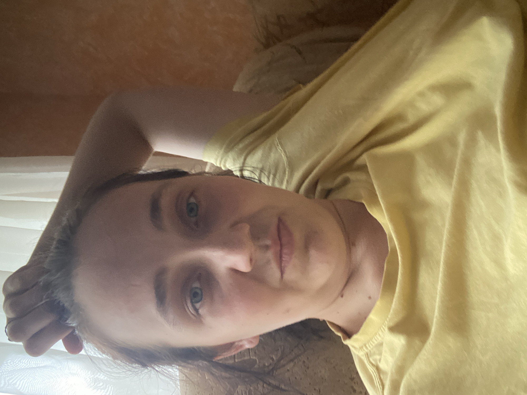 A close-up selfie features a person with fair skin and blue eyes, resting their head on their arm. They are wearing a yellow t-shirt. The background shows a textured wall in an orangey-brown hue and a sheer white curtain. The lighting suggests an indoor setting.