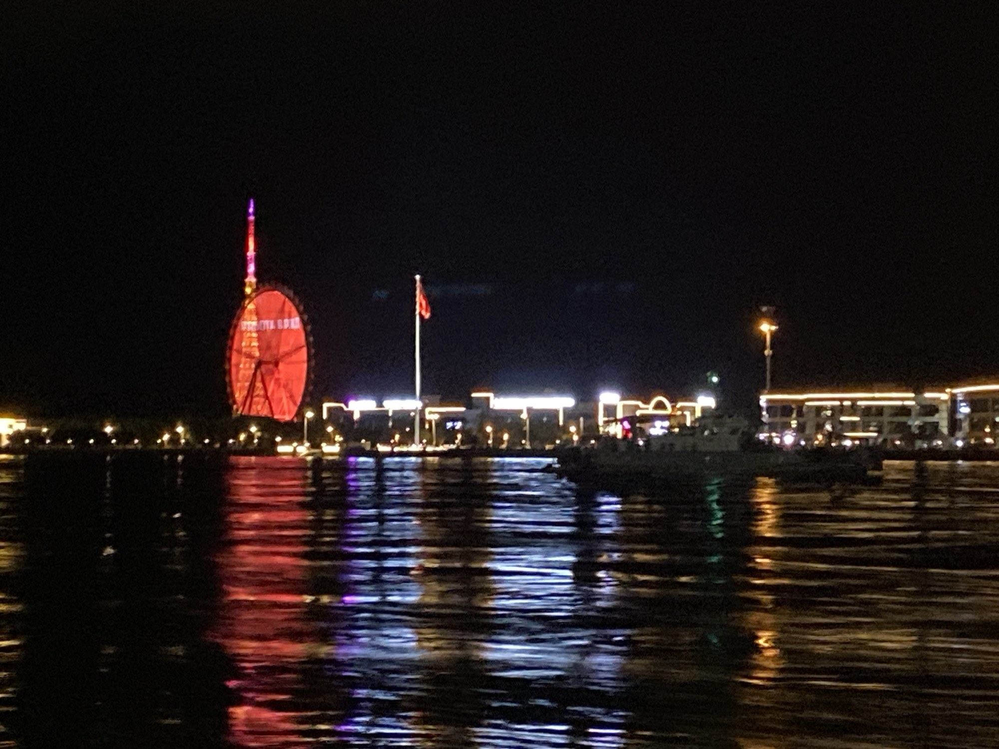 A nighttime, wide-angle shot shows a waterfront scene in Blagoveshchensk, Russia, with a large red Ferris wheel dominating the left side of the frame, illuminated by bright lights. To the right of the Ferris wheel, a flagpole with a red flag is visible against the dark sky, followed by a line of illuminated buildings with windows and decorative lights. A prominent lamppost stands in the mid-right of the image. The foreground is a body of water, likely the Amur River, with its surface reflecting the lights of the city, creating streaks of red, yellow, and blue. A portion of a boat is visible in the water, with its lights also reflected. The overall scene suggests a vibrant, urban nighttime landscape at the river's edge.