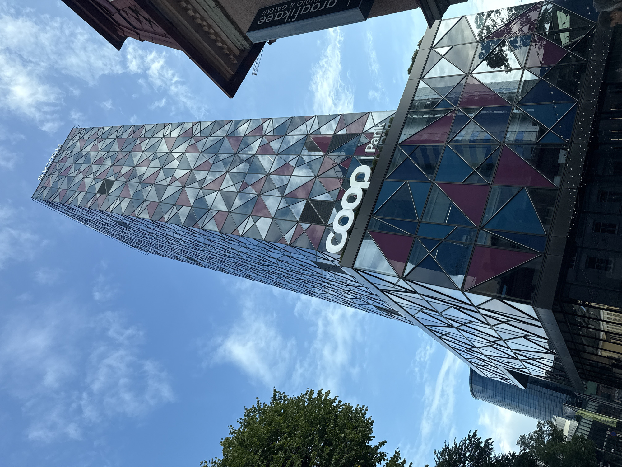 An eye-level, exterior shot captures the distinctive modern architecture of the "Skyon" building, a prominent feature of Tallinn, Estonia's skyline. The building, characterized by its angular, faceted glass facade, displays a dynamic geometric pattern of blue, purple, and mirrored triangular panels. The "coop" logo is prominently displayed in white text on the building's lower section, along with "Park" subtly placed to its right. The structure curves outward towards the bottom of the frame, creating a sense of movement and visual interest.
To the right of the Skyon building, the upper section of a more traditional, older building with a red tiled roof and ornate detailing is visible, creating a stark contrast in architectural styles. A dark, vertical sign with the text "graafika.ee" and "STUDIO & GALLERI" is attached to this older building.
On the left side of the frame, the lush green foliage of a mature tree partially obscures the Skyon building, adding a natural element to the urban scene. In the background, another, sleeker, curved glass skyscraper is visible, suggesting a modern business district. The sky above is a clear blue, dotted with scattered white clouds, indicating fair weather. The perspective is upward-looking, emphasizing the height and grandeur of the Skyon building against the expansive sky. No people are clearly discernible in the image.