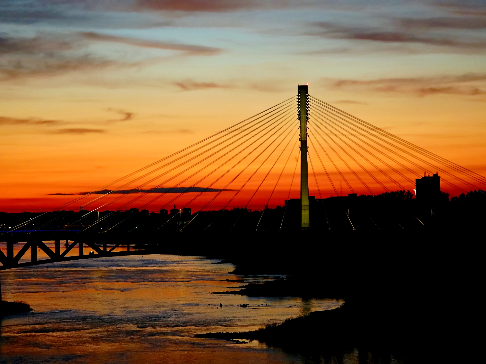 The image captures a dramatic sunset over the Vistula River in Warsaw, Poland. A prominent cable-stayed bridge dominates the right side of the frame, its concrete pylon silhouetted against the vibrant orange and red hues of the sky. Numerous cables fan out from the pylon, supporting the bridge deck, which is also silhouetted. A smaller, more traditional truss bridge is visible on the left side of the image, partially obscured by the river's reflections. The river itself reflects the fiery colors of the sunset, creating a shimmering, fluid surface. The city skyline is a dark silhouette in the background, punctuated by the faint lights of buildings and streetlamps. The overall impression is one of urban grandeur set against a spectacular natural backdrop. There are no visible people or animals in the image.