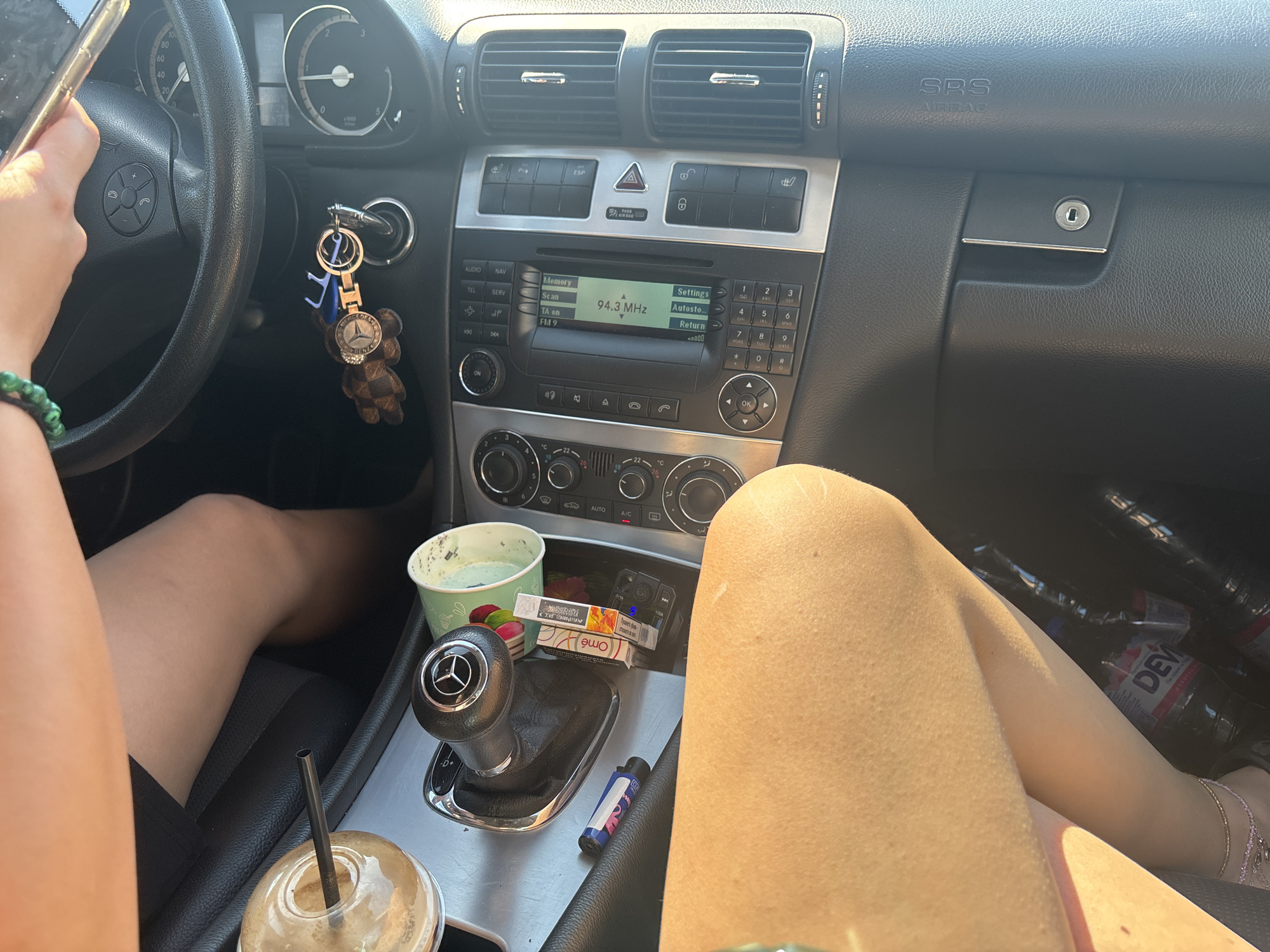 A passenger's legs are visible in the foreground of the image, which appears to be taken inside a car in Sofia, Bulgaria. The passenger is wearing shorts. The dashboard and center console of the car are also visible. On the center console, there is a gear shift lever with a Mercedes-Benz logo, a cup of coffee, cigarettes, a lighter, and a car key with a keychain. The car radio is displaying "94.3 MHz". The overall scene suggests a casual setting, possibly during a road trip or a stop in the city.