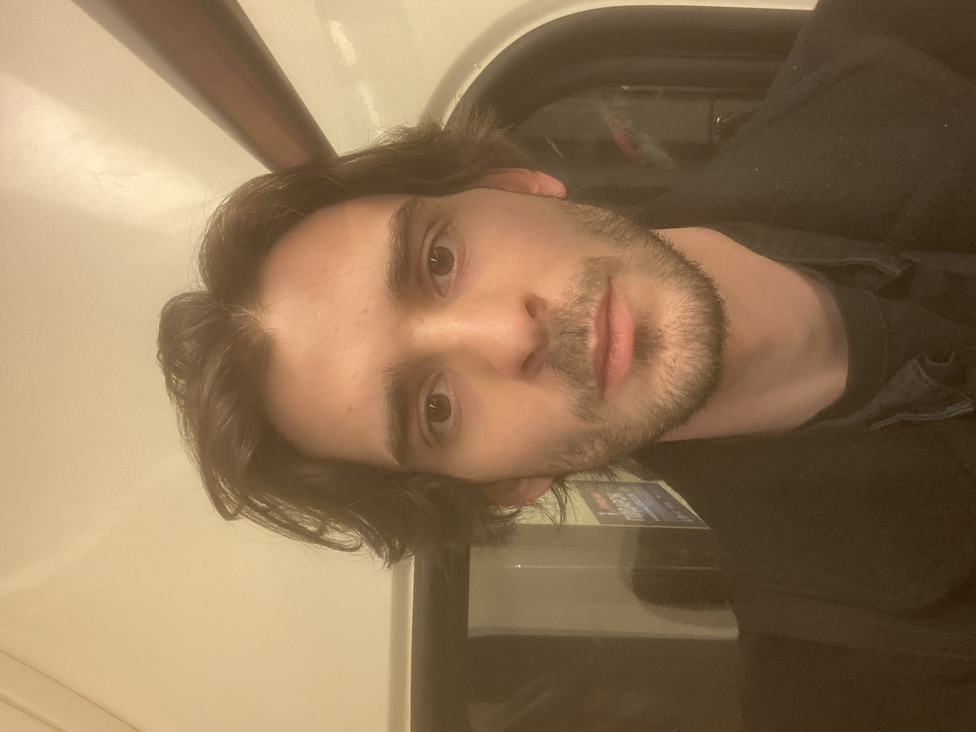 A young man is sitting inside a train, presumably a subway car. The train car appears to be fairly standard, with a long interior. The train seems to be in motion. The man has dark hair, stubble, and is wearing a dark jacket. The location cannot be determined from the image.