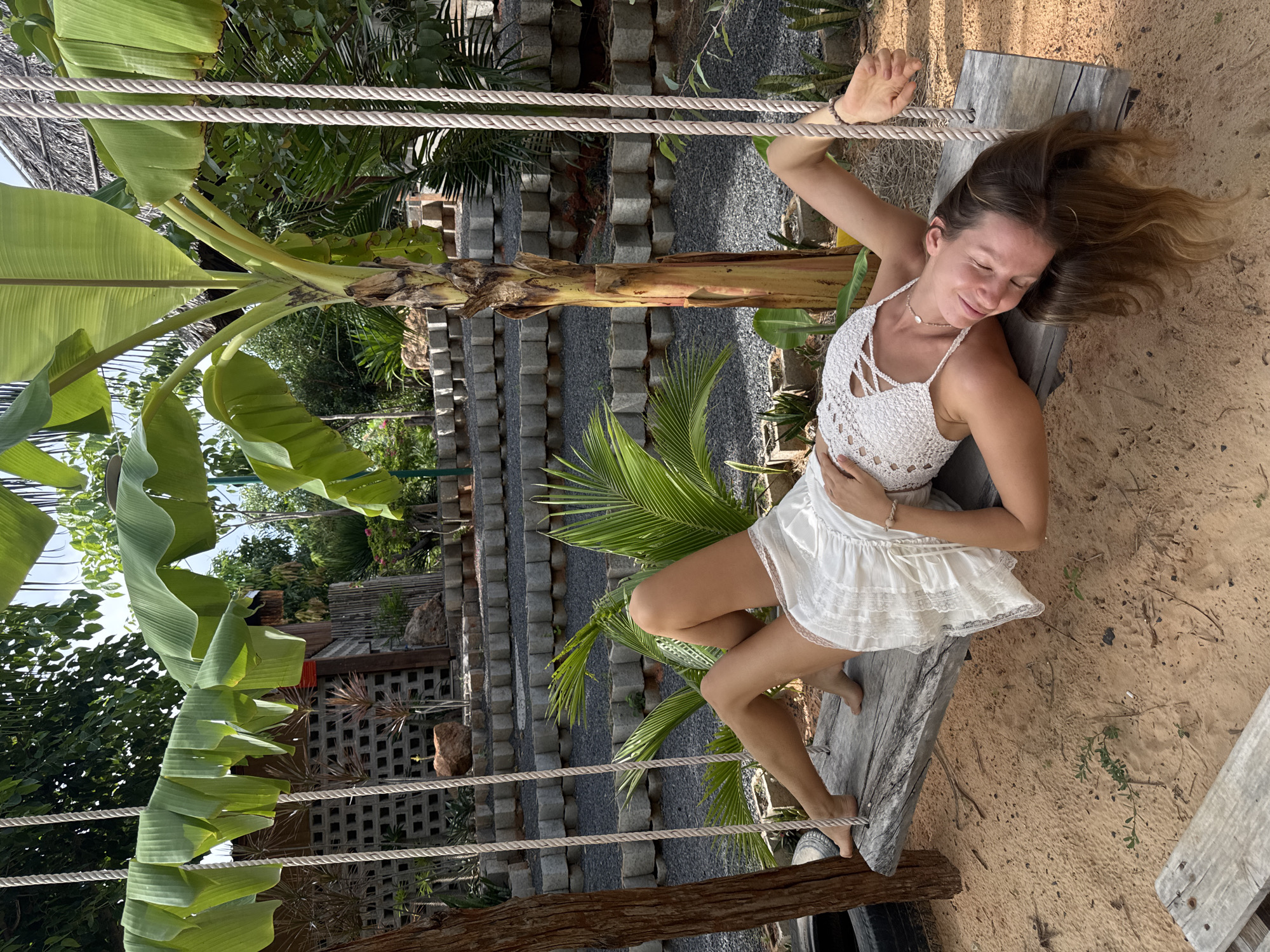 A woman in a white dress and top is lying on a wooden swing with her eyes closed. She is positioned on the right side of the swing, with her head resting on the left end. Her left arm is bent at the elbow and resting behind her head, while her right arm is extended to her side. Her legs are crossed at the ankles and extended upwards. The swing is suspended by ropes from a tree and is set against a backdrop of a sandy ground, tropical foliage, and a tiered stone structure. A banana tree is prominently visible behind the swing, its large leaves framing the scene. The location is identified as Mũi Né, Vietnam, suggesting a tropical or coastal environment.