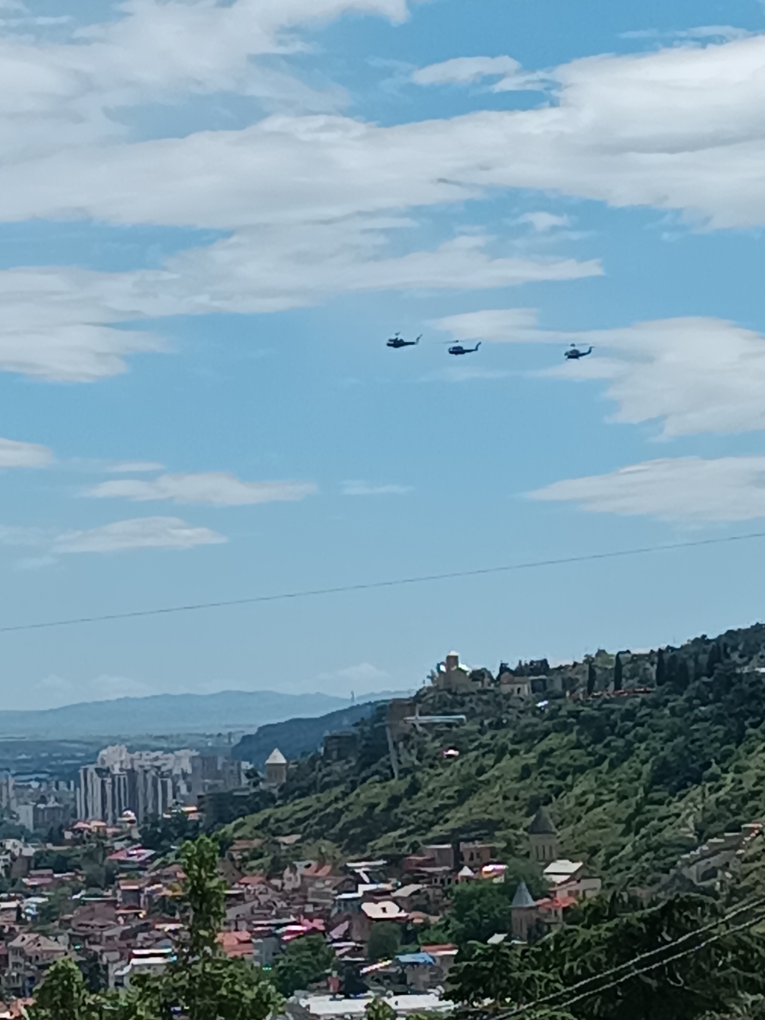 The image shows the city of Tbilisi, Georgia, with a hilltop featuring historical structures. In the foreground, there are red-roofed buildings and green trees. In the distance, there is a cluster of modern high-rise buildings and hills. Three helicopters fly in formation across the blue sky dotted with white clouds.