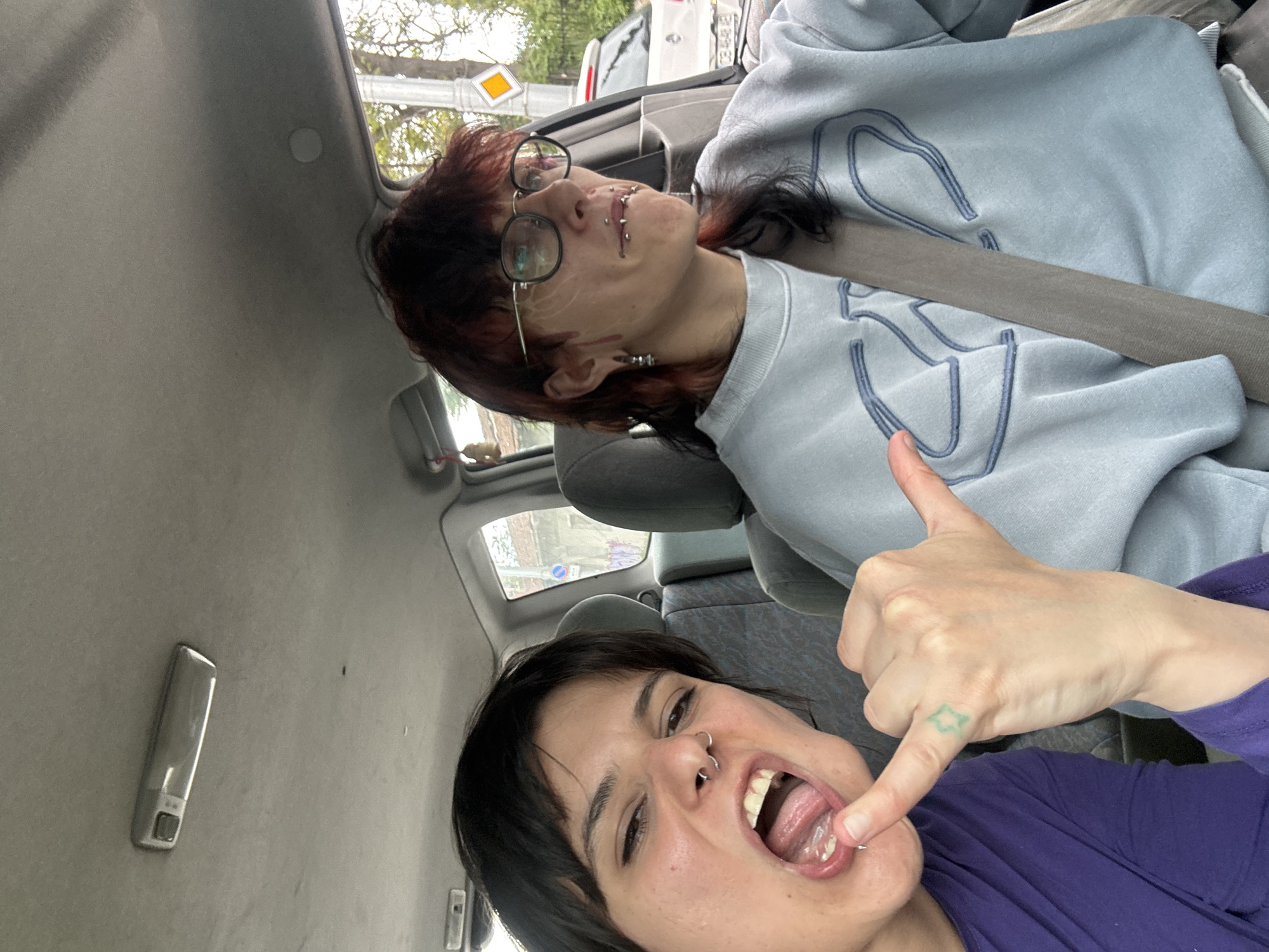 Two individuals are inside a vehicle. The person in the foreground is making a face with their mouth open, and a visible tongue piercing. They are wearing a purple top, and there is a tattoo visible on their hand. The person in the background is wearing glasses and a light blue sweatshirt. They have multiple facial piercings and a dark-colored ponytail. Both individuals appear to be wearing seatbelts. Outside the vehicle, a street sign is visible. Based on the street sign's appearance, it is possible this image was taken in Sofia, Bulgaria.
