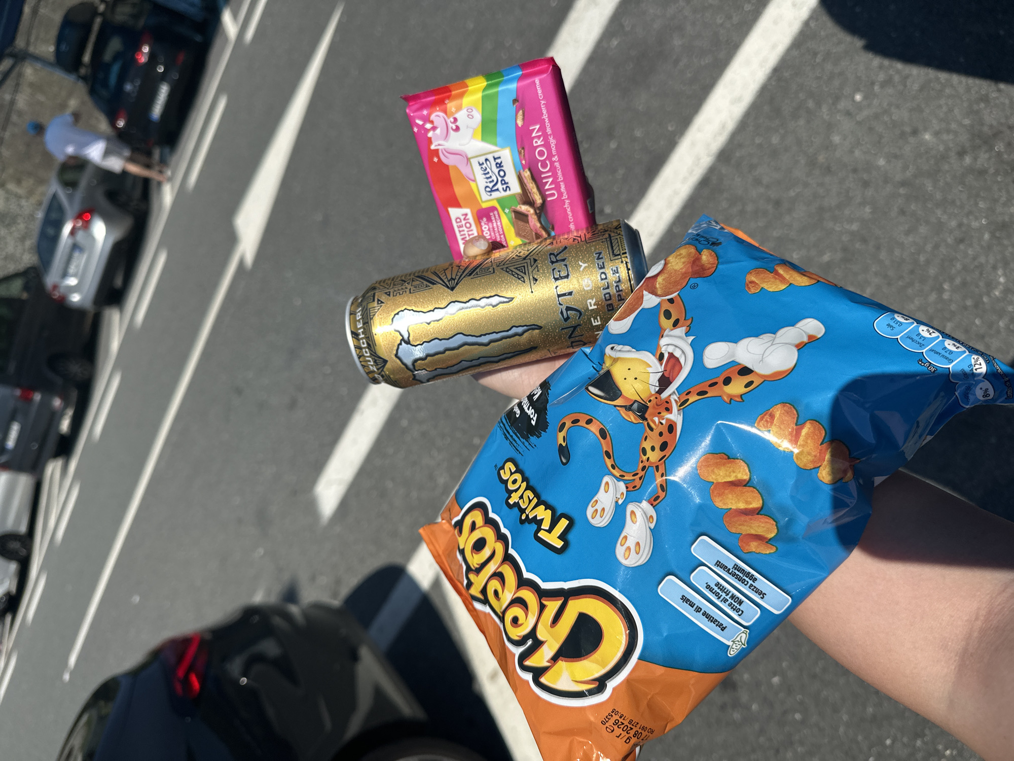 The image shows a person's hand holding a bag of Cheetos Twistos, a can of Monster Energy Golden Apple, and a Ritter Sport Unicorn chocolate bar. The background is a street with parked cars and white lane markings, suggesting a parking area or roadside. The location appears to be in Italy based on the Italian text on the Cheetos package, specifically "Patatine di mais," "Senza conservanti," "Cotte al forno, NON fritte." It cannot be definitively ascertained from the image that the location is Varazze, Italy.