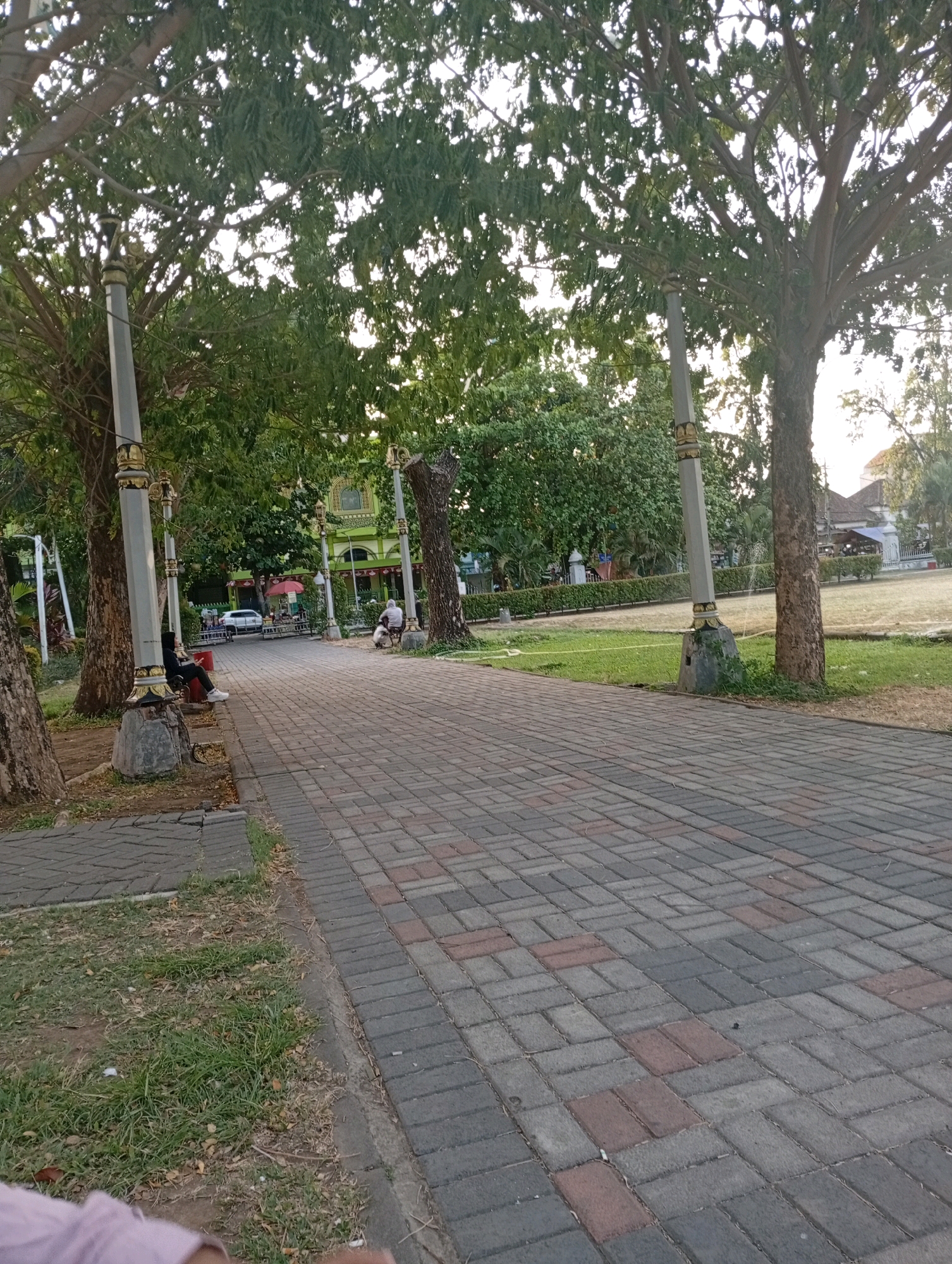 The image depicts a paved pathway in a park-like setting, likely in Bangil, Indonesia. The pathway is constructed from rectangular bricks in various shades of grey and red, arranged in a herringbone pattern. Tall trees with dense green foliage line the pathway, casting shadows and dappling sunlight. Lampposts with decorative gold accents are strategically placed along the path, suggesting it is a public area.
In the background, a light green building with arched windows and a tiled roof is visible, possibly a mosque or another significant public structure. To the right of the pathway, a grassy area stretches out, with more trees and some fencing.
Several people are present in the scene. On the left side of the pathway, a person is sitting on a stone bench, wearing dark clothing and white shoes, looking towards the right. Further down the path, another individual appears to be riding a bicycle, also facing away from the camera. In the distance, near the green building, a white car is parked. A red umbrella is also visible near the car, suggesting a small vendor or seating area. The overall atmosphere is peaceful and suggests a typical afternoon in a community park.