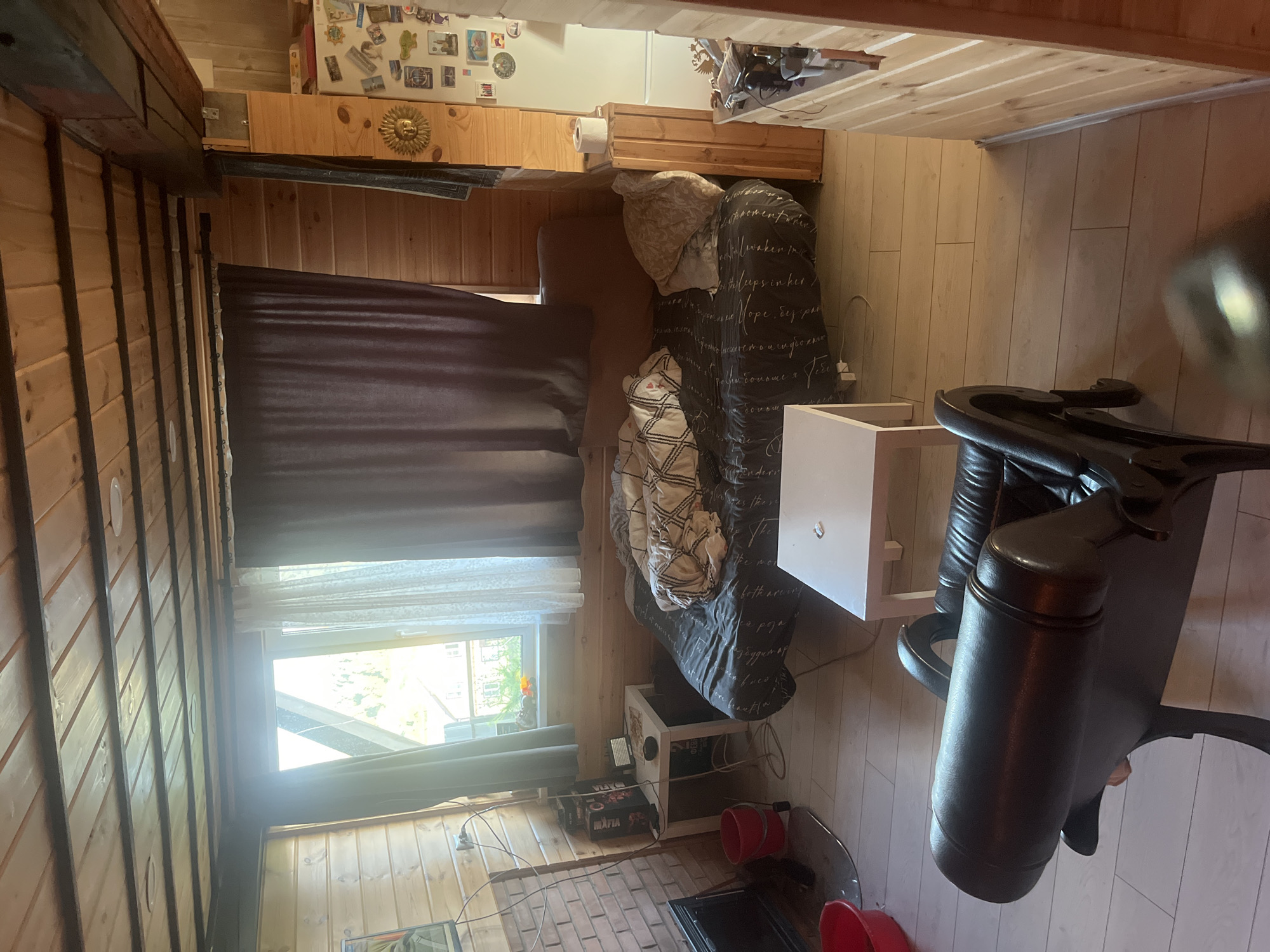 The image shows an interior view of a room with wooden walls and ceiling. The room appears to be a living space or bedroom with a couch covered in a dark patterned duvet and a blanket. There are several pillows on the couch. To the left of the couch, there is a small white table with some items on it, including a box and a device. A fireplace is visible to the far left. In the foreground, a dark leather chair is partially visible. A window with sheer curtains and dark drapes is to the left of the couch. To the right of the couch, there is a white cube-shaped table. Further to the right, a refrigerator is adorned with various magnets and pictures. The floor is made of light-colored wooden planks. The overall impression is of a cozy, somewhat cluttered, rustic interior. The location is identified as Satka, Russia, suggesting this is a room within a dwelling in that city.