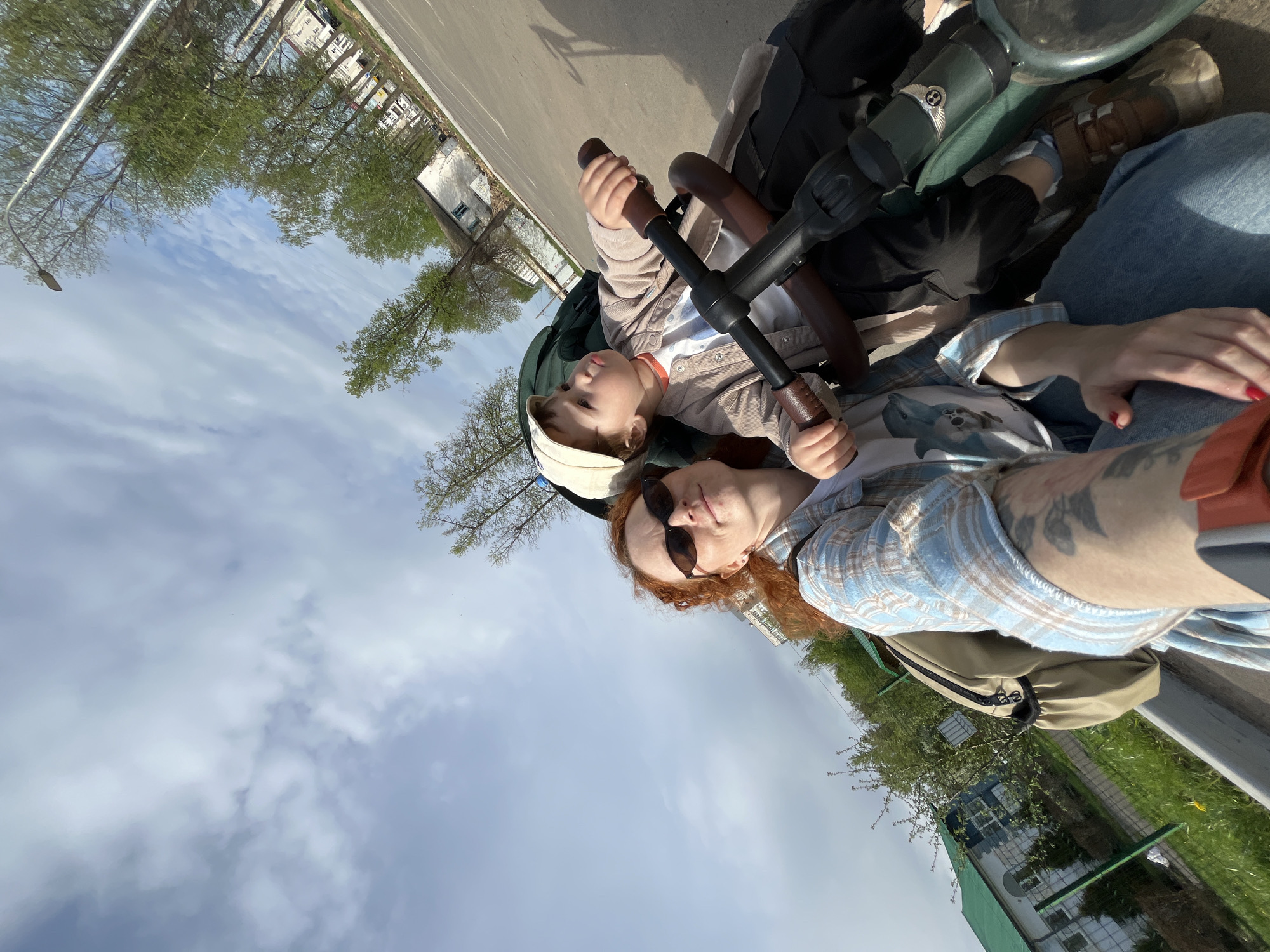 A woman with red hair and sunglasses and a young child are in a stroller on a city street in Chaikovsky, Russia. The woman is wearing a blue and tan plaid shirt and jeans. The child is wearing a white cap and a tan jacket. The stroller is black and green. The street is asphalt, with buildings and trees in the background. The sky is blue with white clouds.
