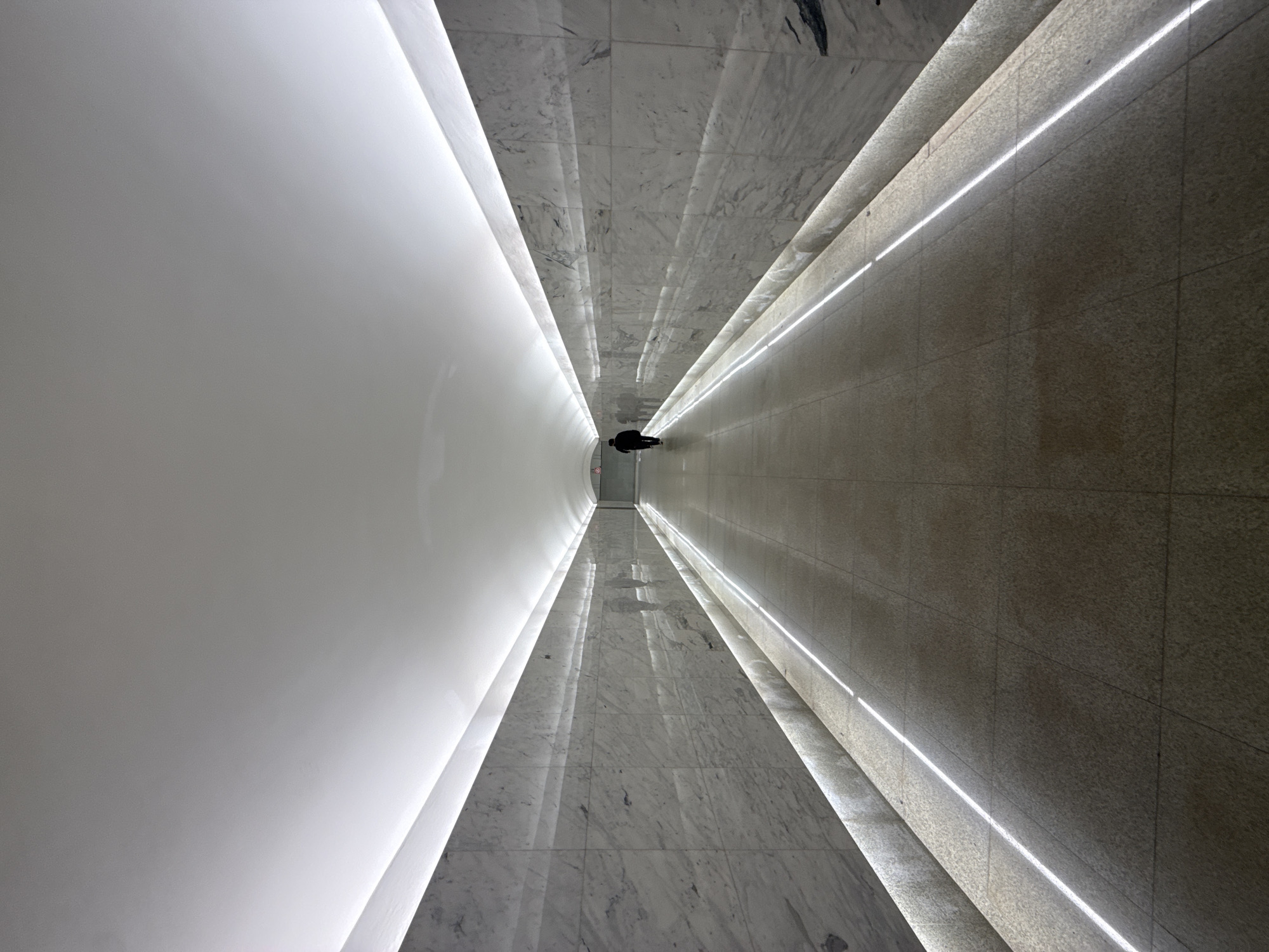 The photograph shows a long, brightly lit corridor from an overhead perspective. At the end of the corridor, a person is walking away from the viewer. The left wall is plain white, while the floor and right wall are covered in marble. Linear lighting runs along the floor and right wall. Based on the location, this is likely a hallway at the Moscow International Business Center in Moscow, Russia.